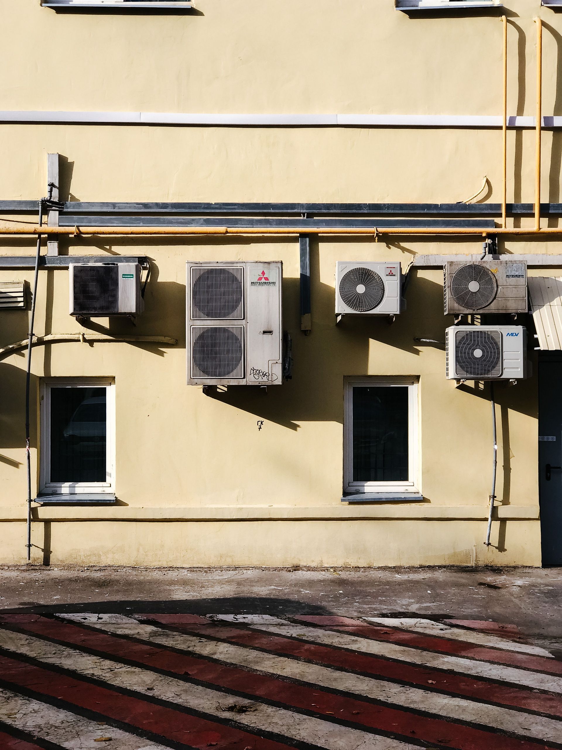 Beige building wall with windows, air conditioners, and red-striped pavement below