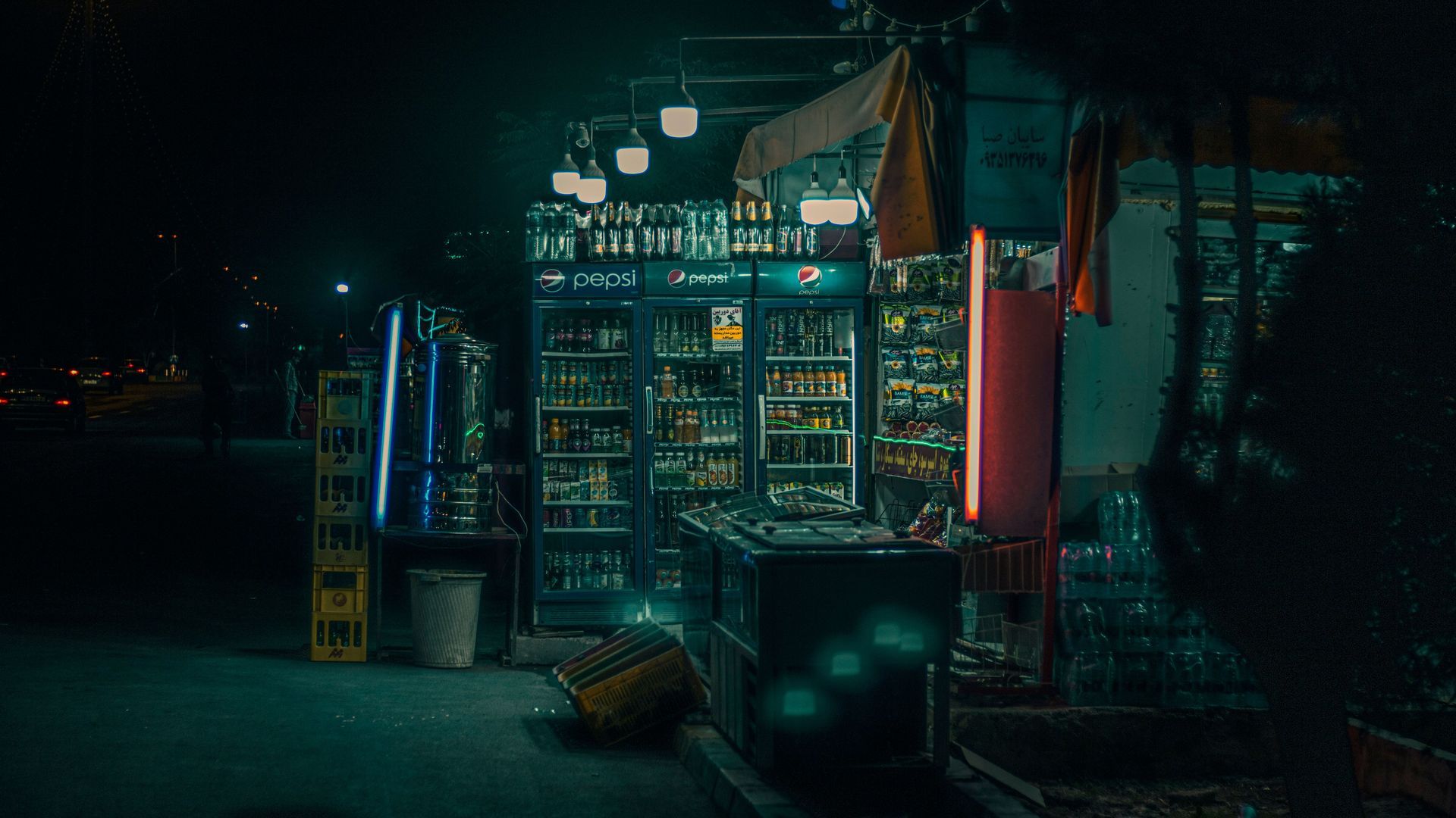 Dimly lit nighttime street kiosk with glowing signs and bottled goods in a narrow alley