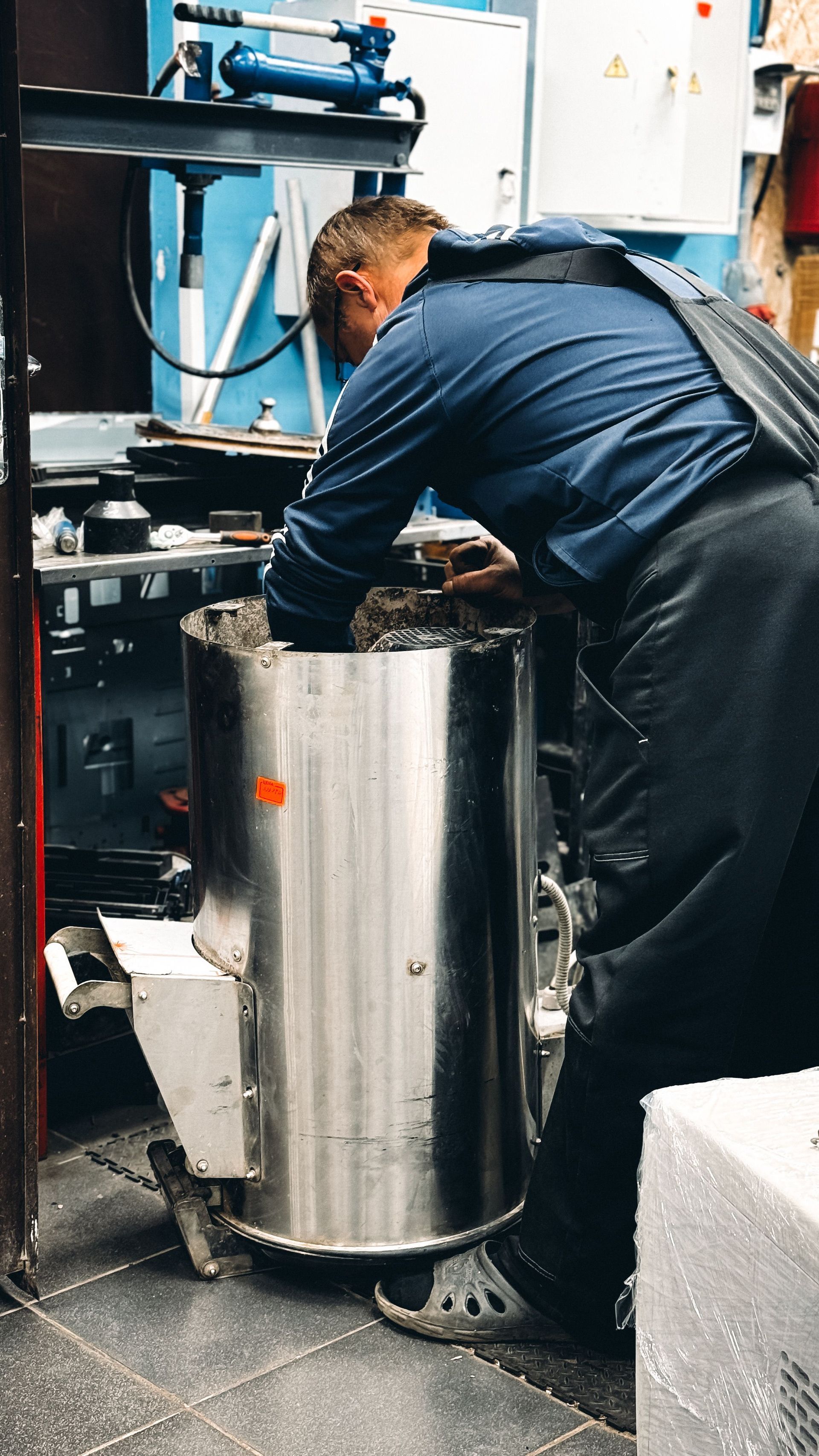 Worker in dark coveralls adjusts machinery beside a large stainless steel tank in an industrial setting