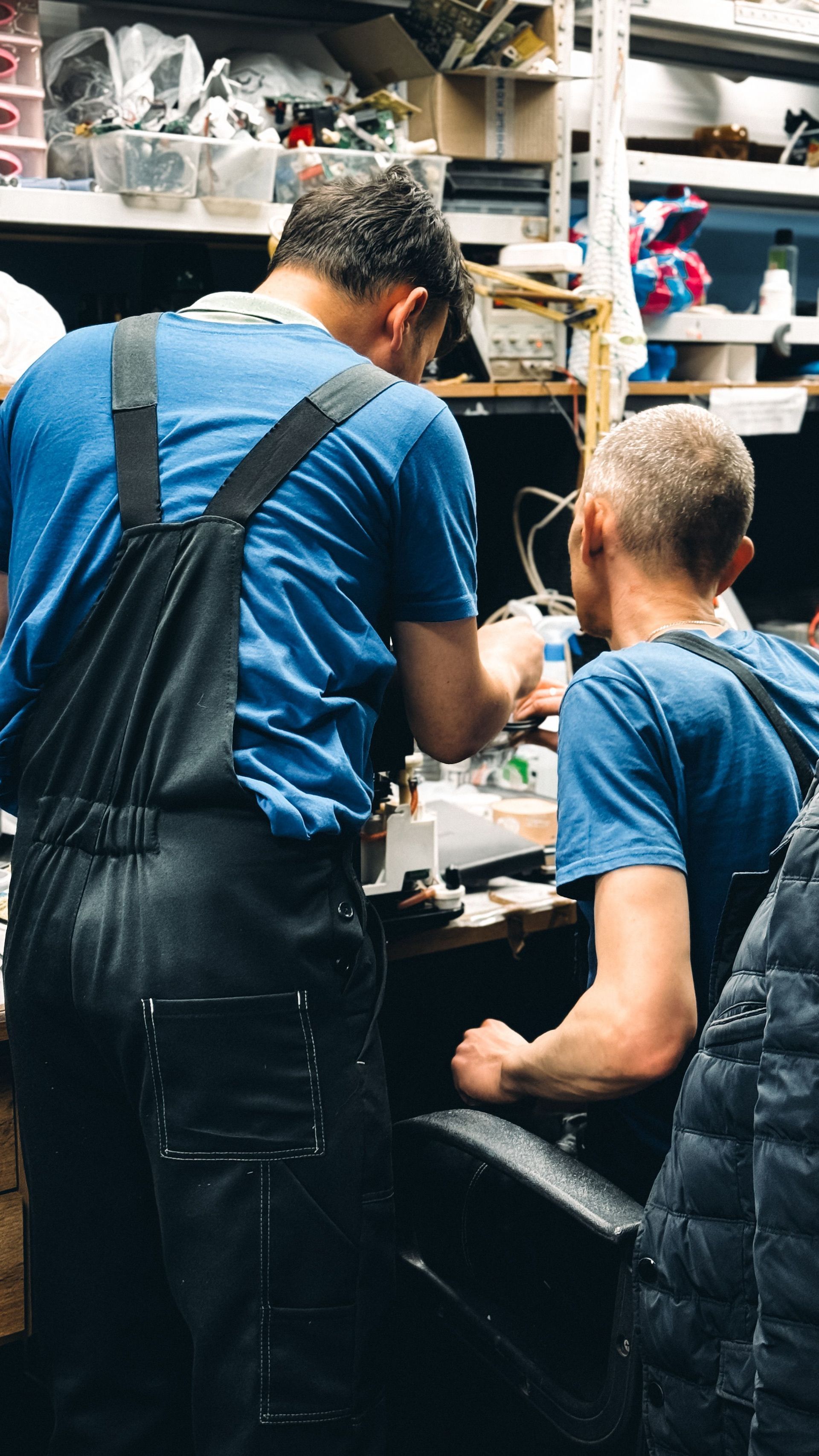 Two people in blue shirts working together in a cluttered workshop, seen from behind.