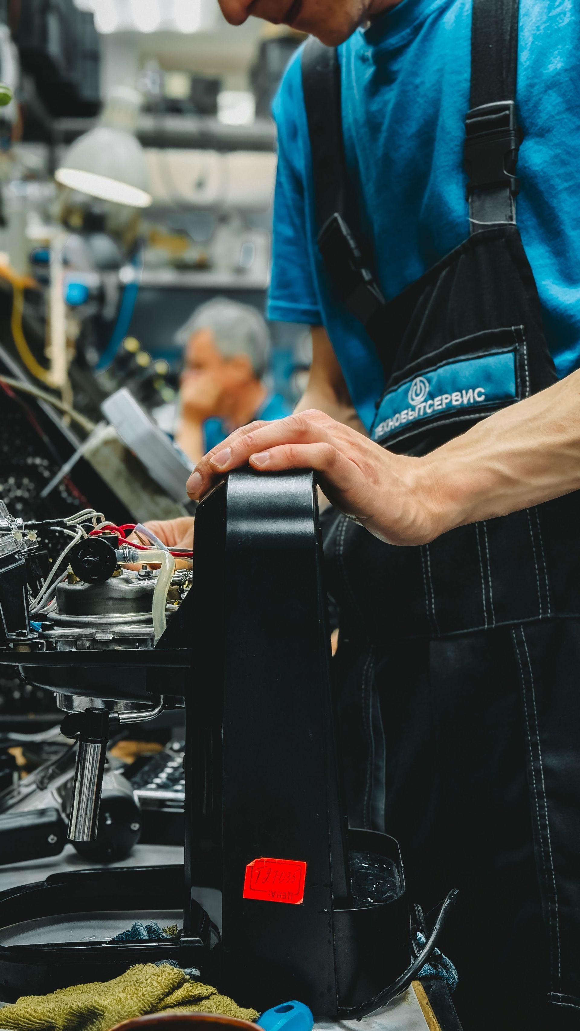 Factory worker handling a black shoe on an assembly line, with machinery and blue-clad workers in the background
