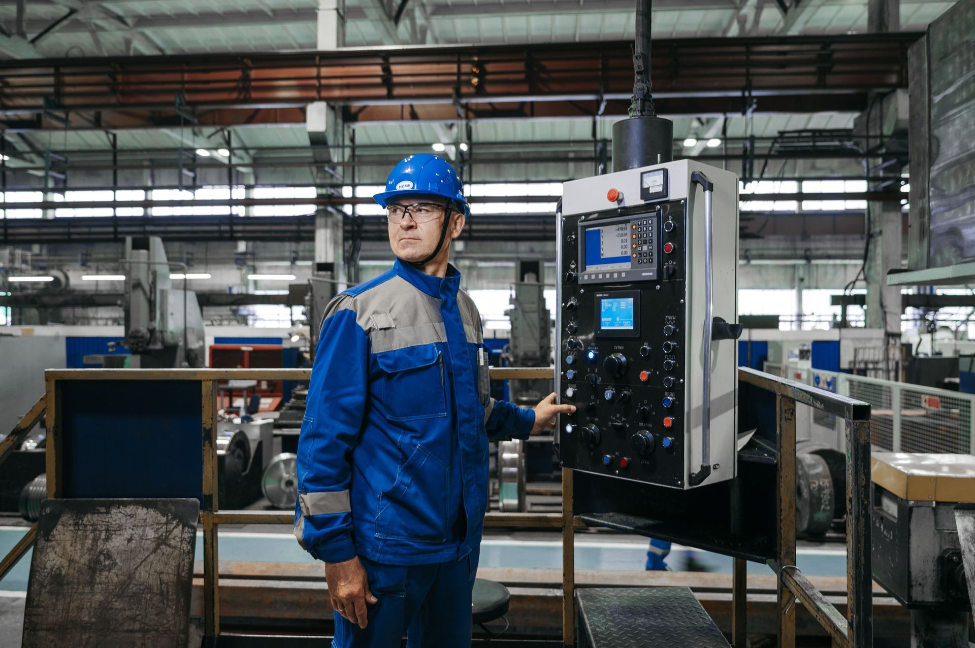 Factory worker in blue uniform and hard hat beside a machine control panel in an industrial workshop