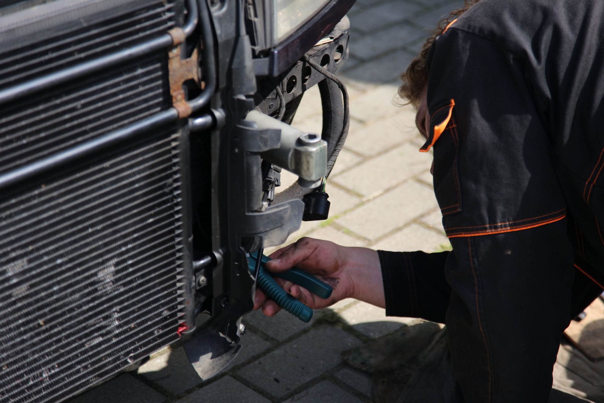 Technician using pliers to work on a car’s front radiator in a driveway