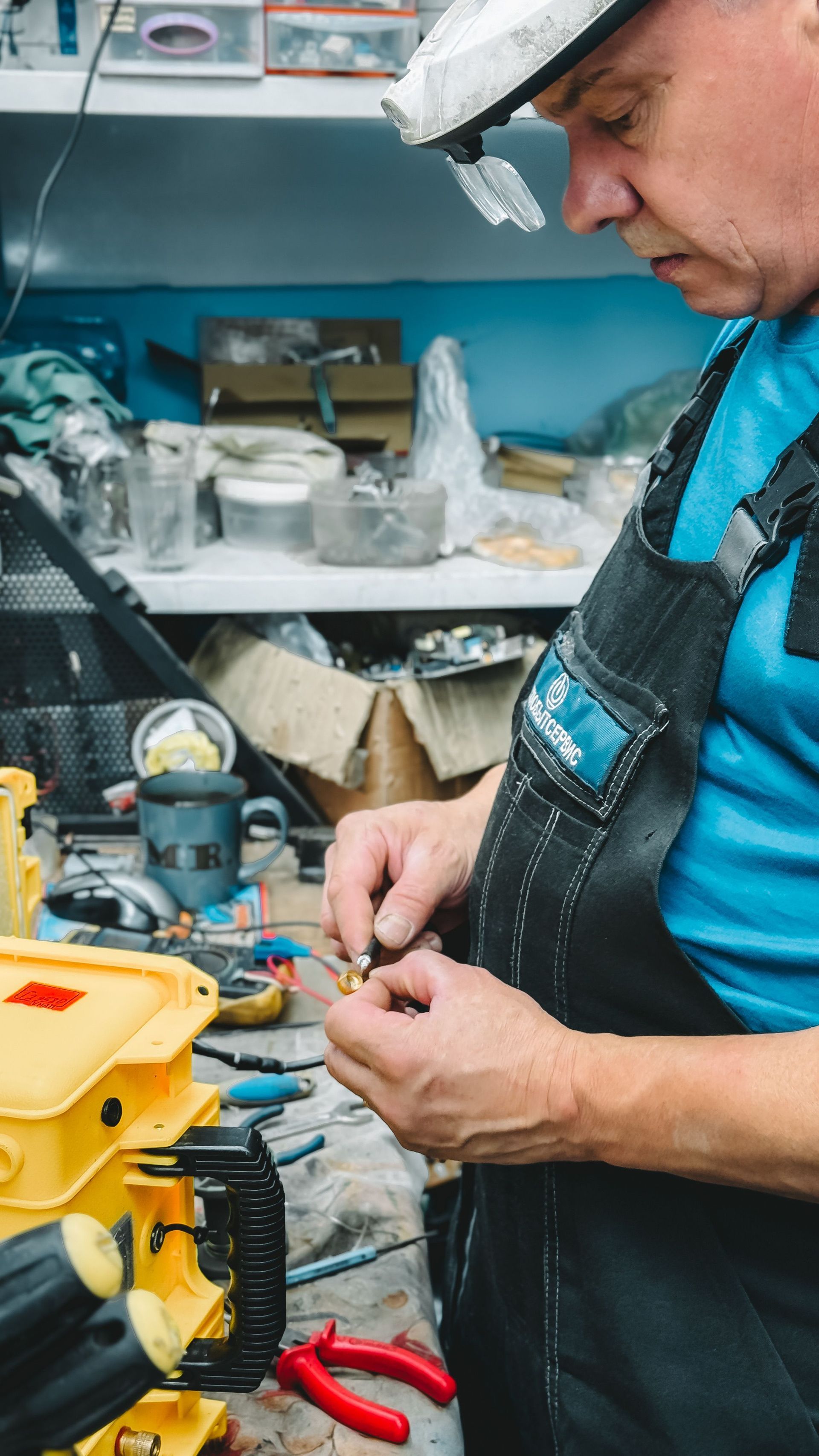 Technician in a blue shirt and black vest adjusts tools at a cluttered workbench with a yellow machine.