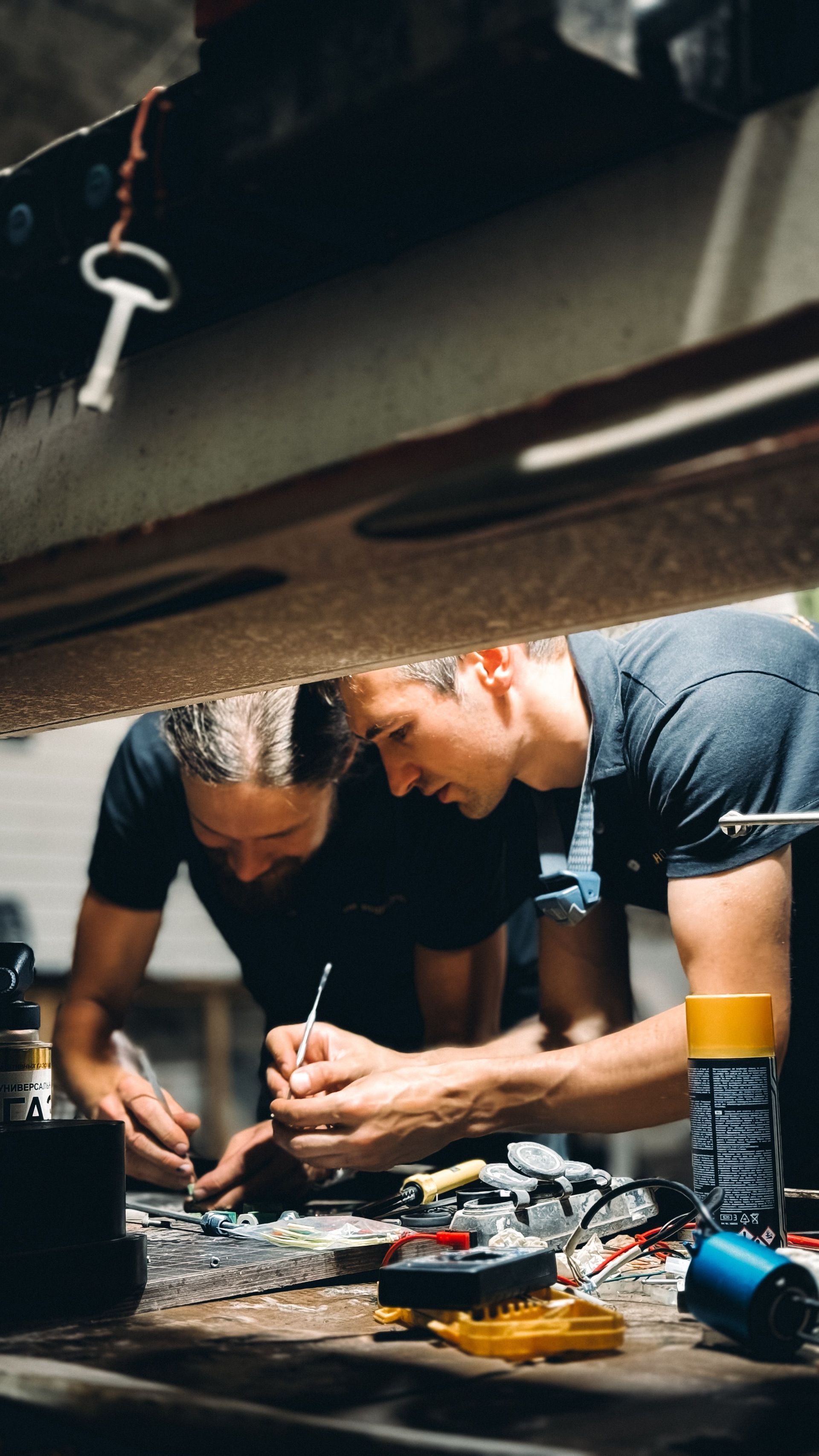 Two workers assembling electronics on a workbench under a car, with tools and wires scattered around.
