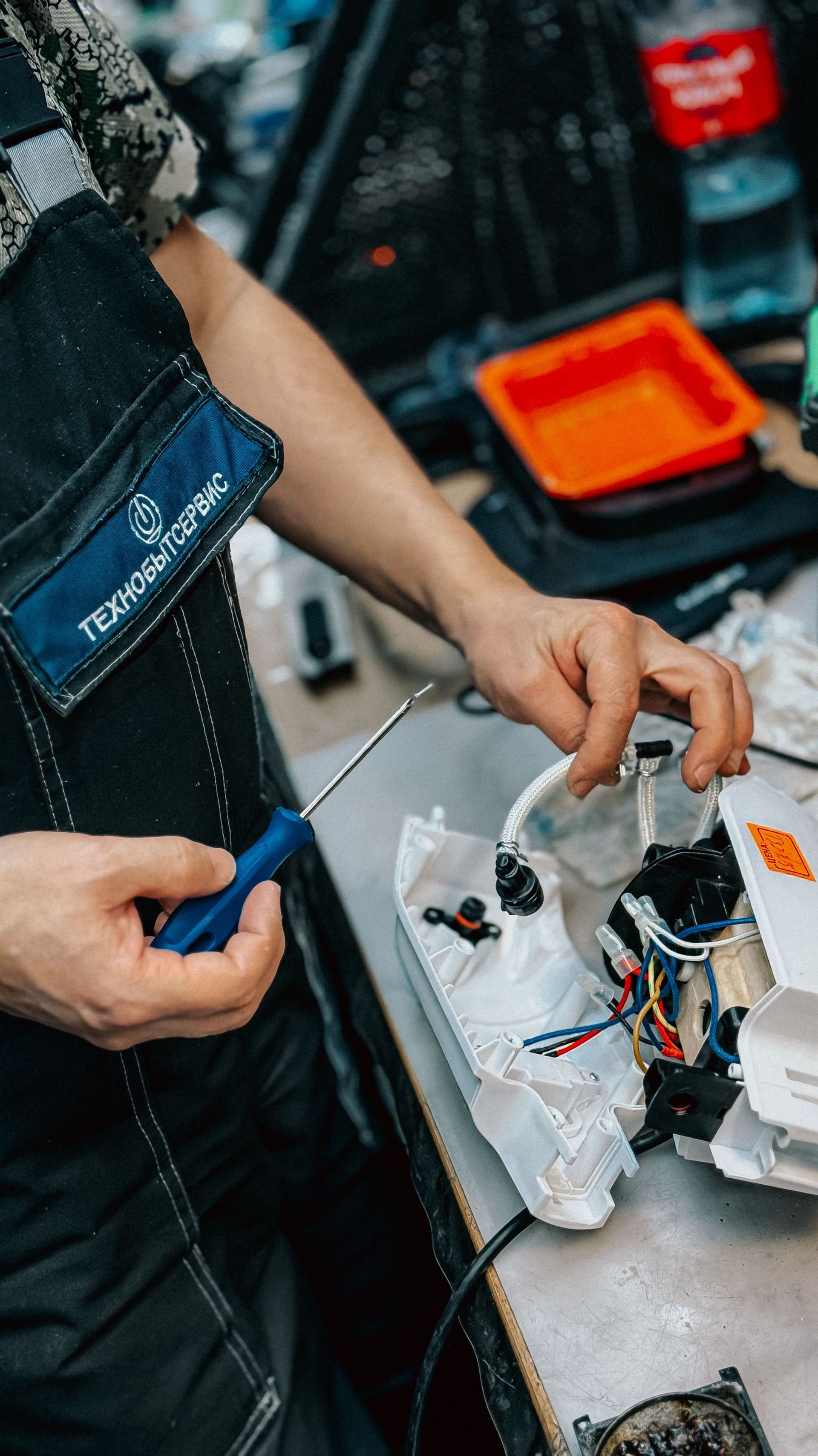 Person holding a blue tool over a messy workbench with wires, containers, and a metal device