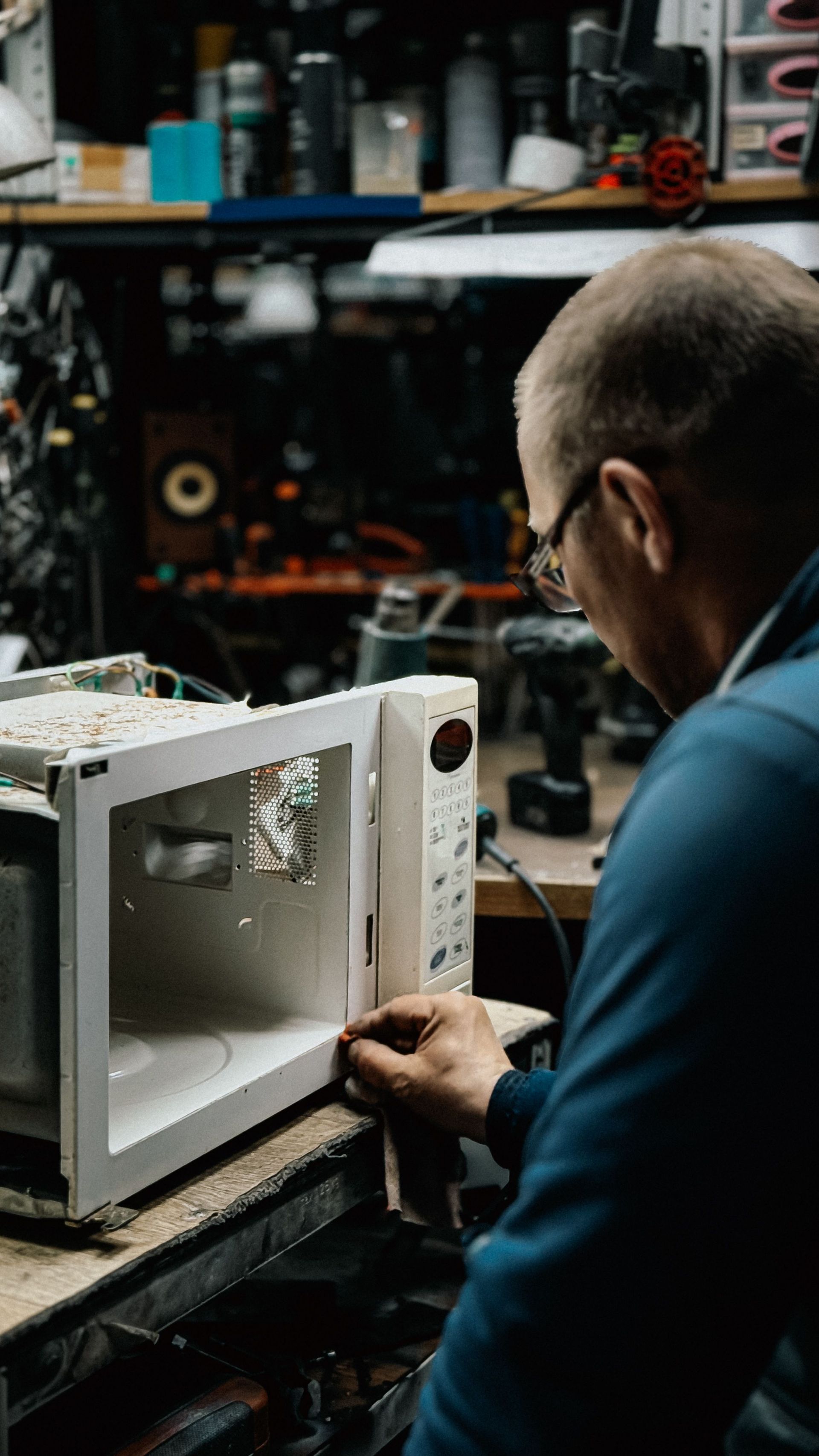 Technician inspecting a microwave in a cluttered workshop, with tools and equipment in the background