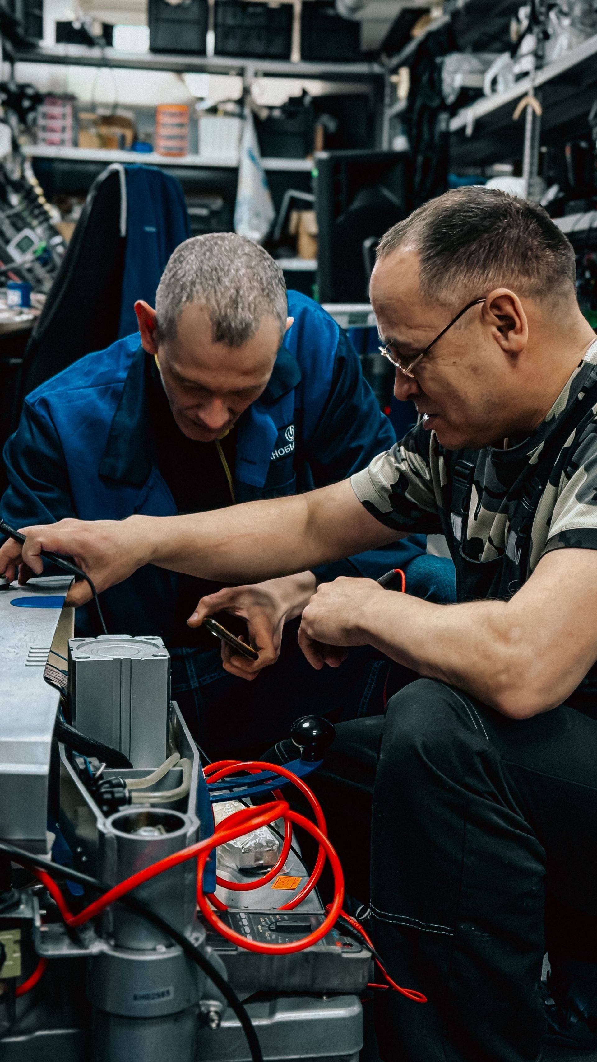 Two men crouch over a small device in a workshop, working with tools and cables.