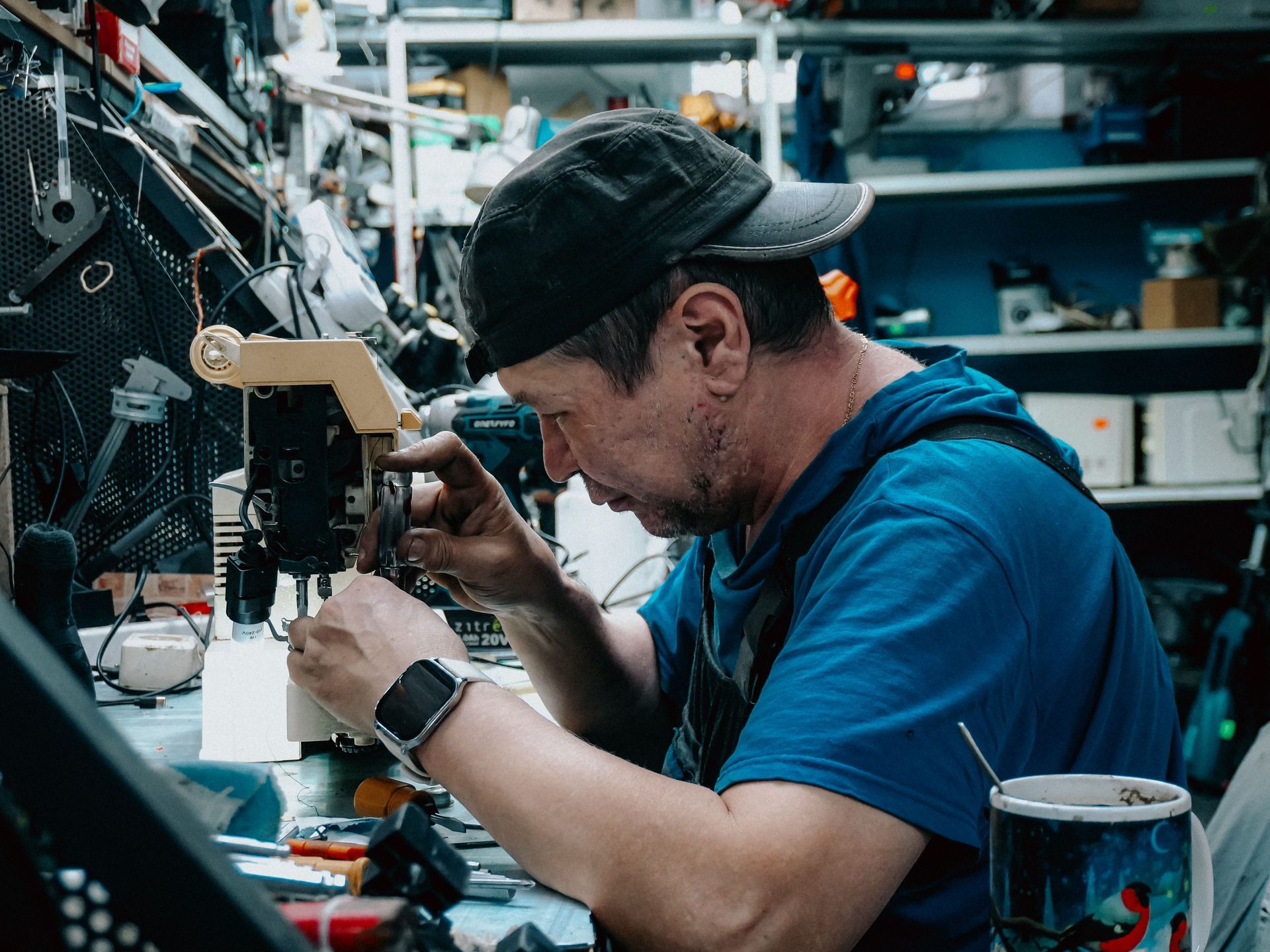 Technician in a blue shirt soldering electronics at a cluttered workbench in a workshop