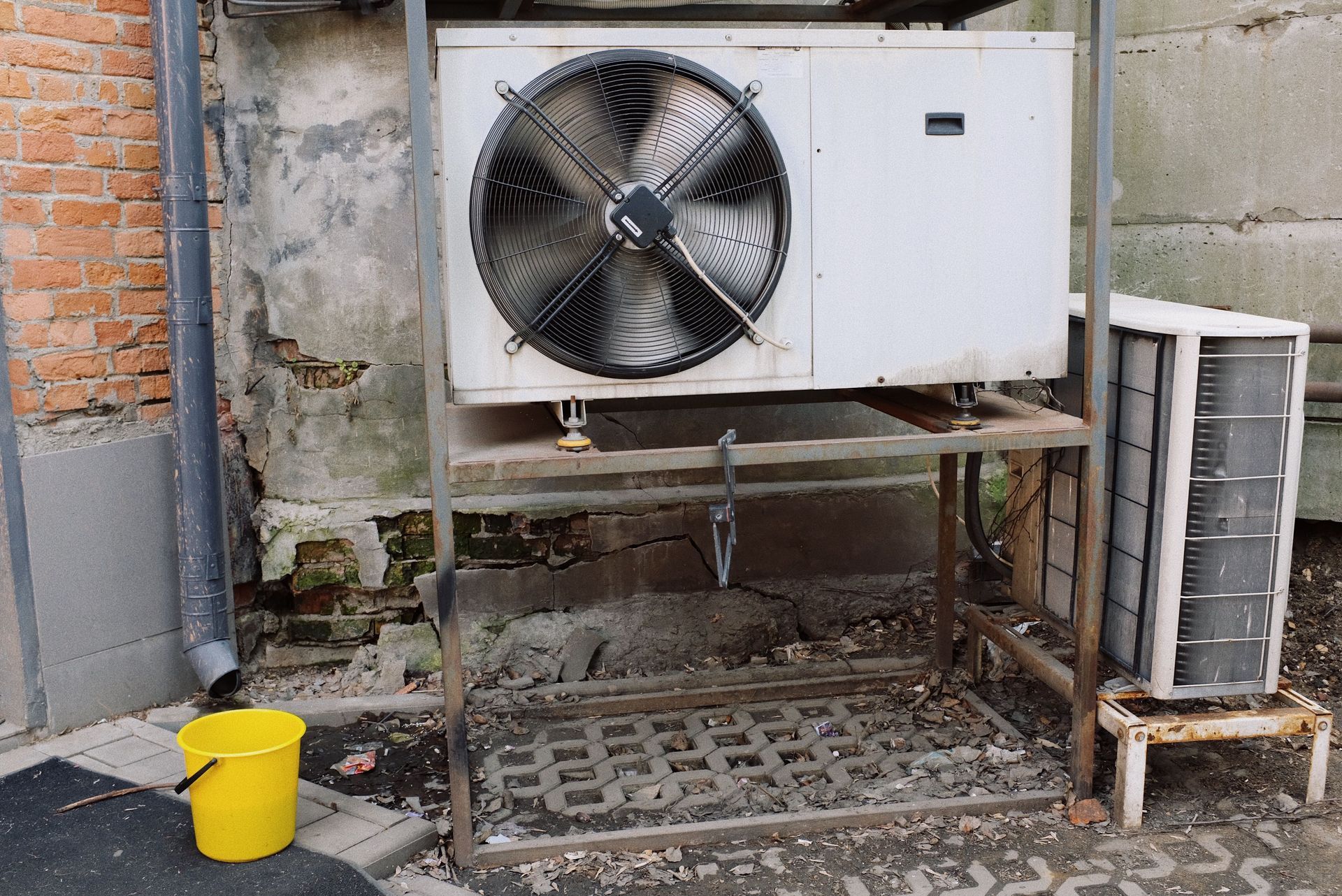 Outdoor air conditioner unit with a large fan beside a brick wall, with a yellow bucket and stacked AC coils nearby