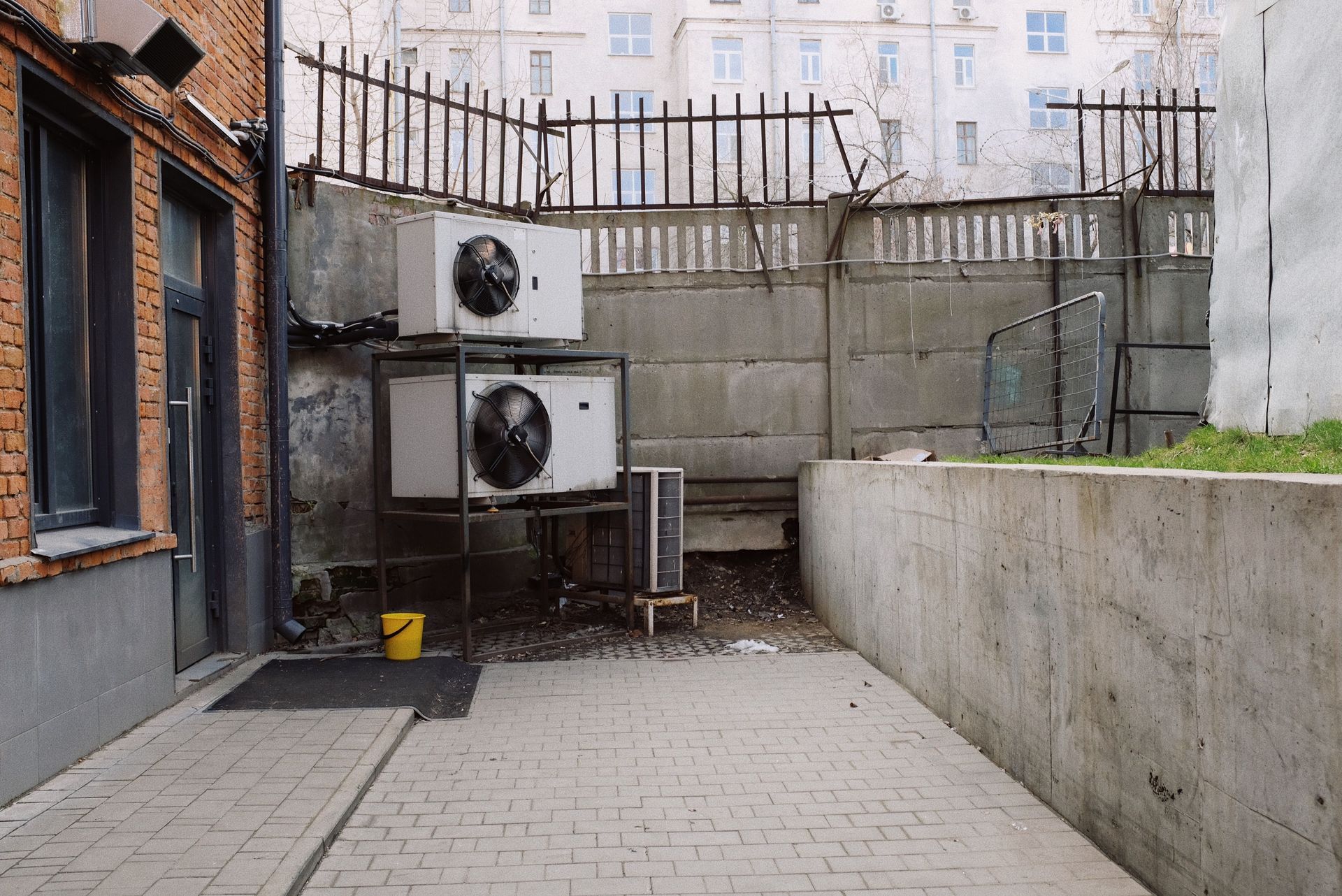Narrow outdoor courtyard with brick paving, concrete walls, and two air-conditioning units against a wall.