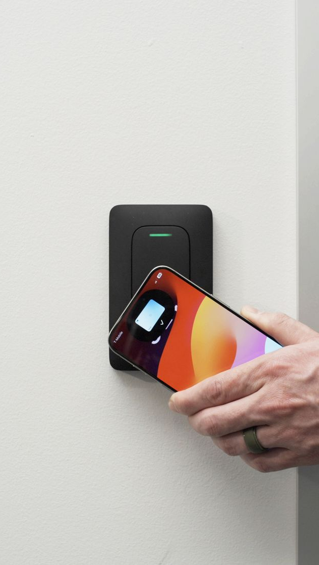 Hand holding phone near a black door access reader on a white wall. Green light on.