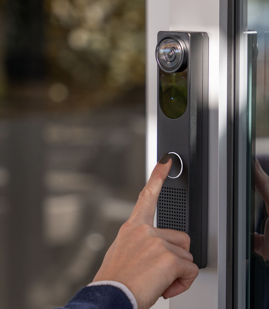 Person's finger pressing a doorbell button on a sleek, modern, black access panel next to a glass door.