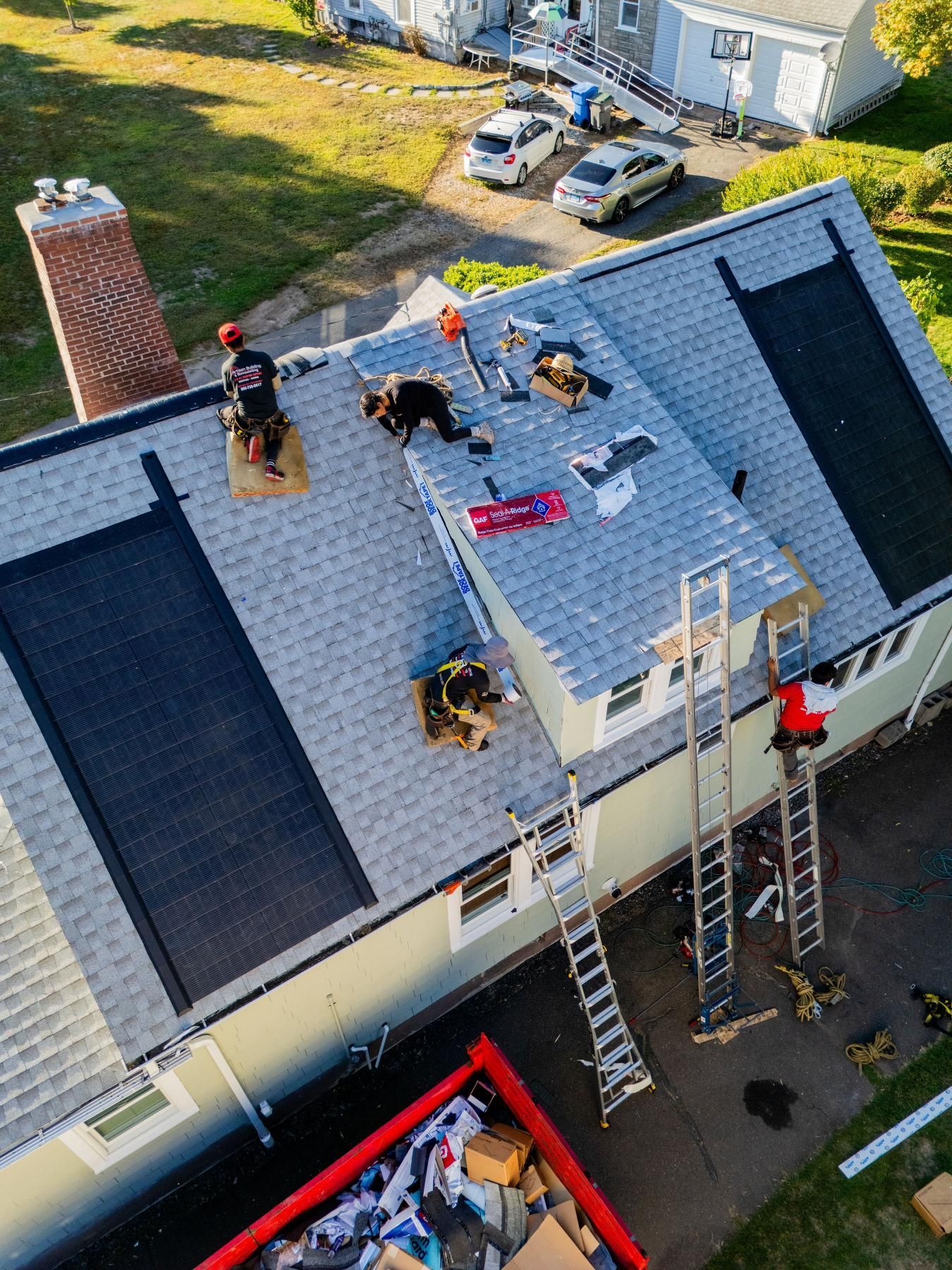 Roofers working on a house roof with solar panels, ladders, and a dumpster in a residential area.