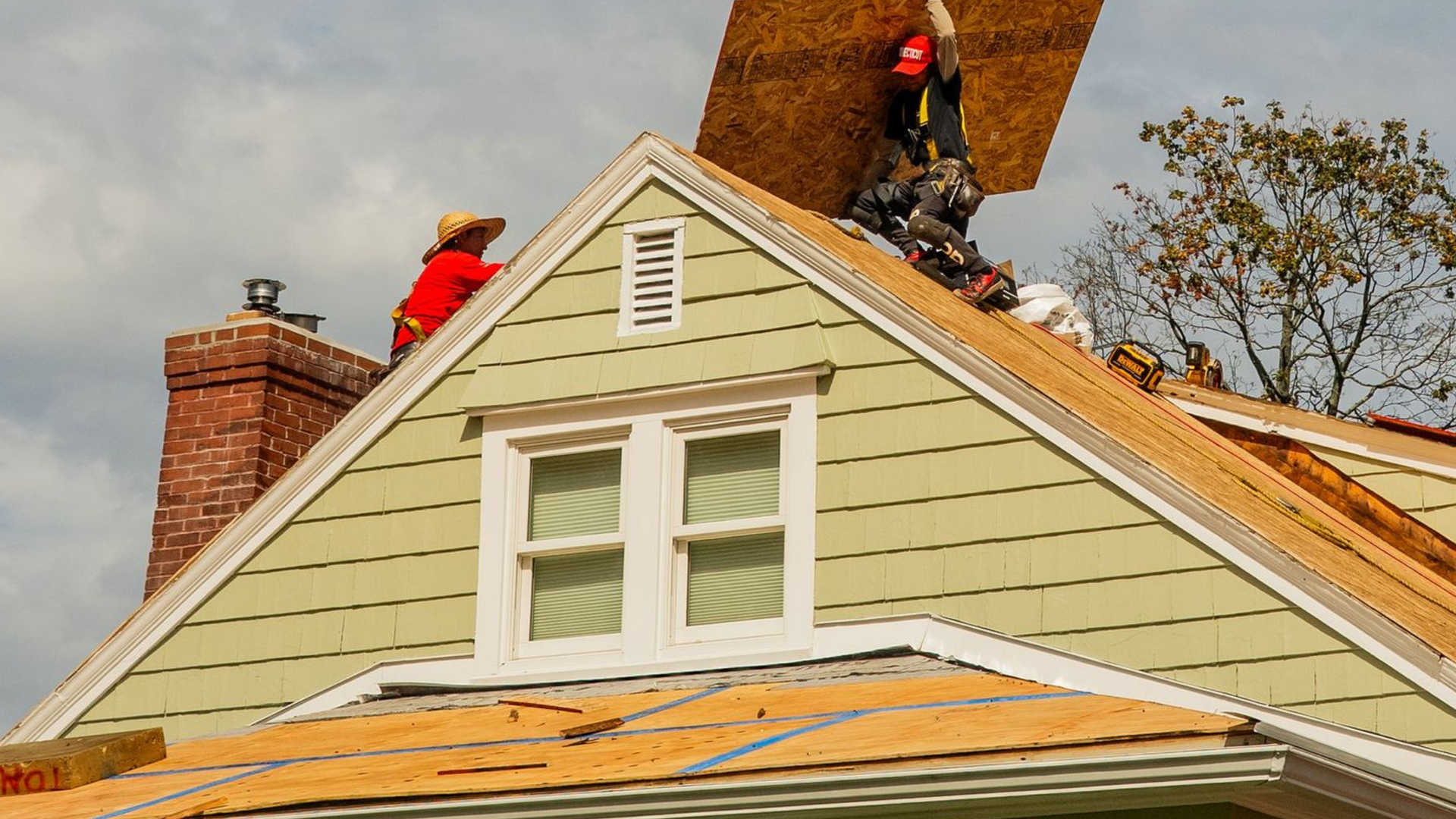 Roofers installing wood sheathing on a gable roof; one in red, another holding up the board.