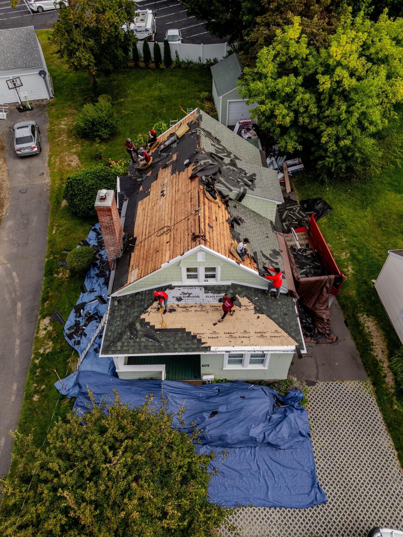 Roofers working on a house roof replacement. Blue tarp covers the ground, with a dumpster alongside the house.