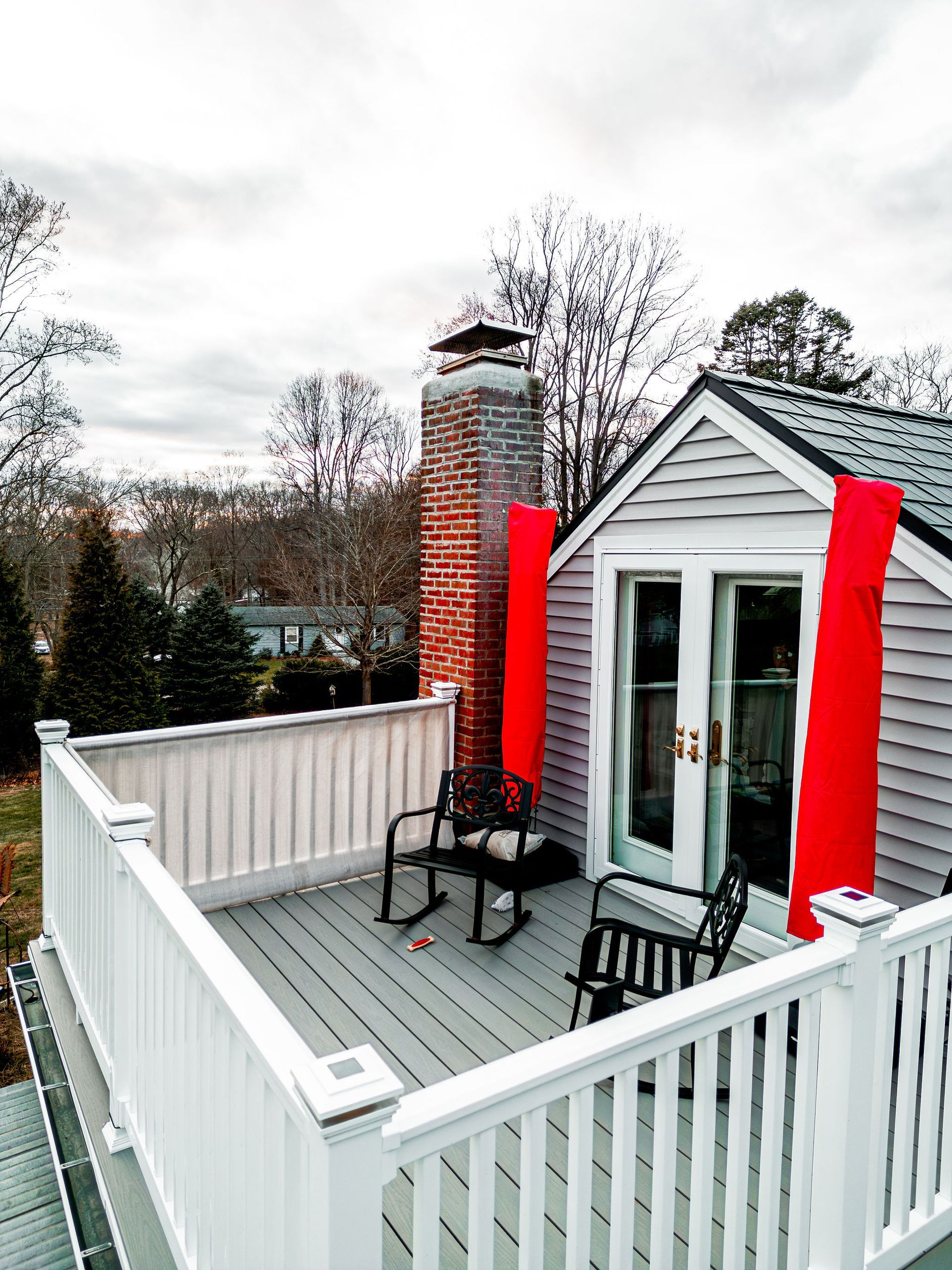 Rooftop deck with gray planks, white railing, and two black rocking chairs. Red accents flank the door and chimney.