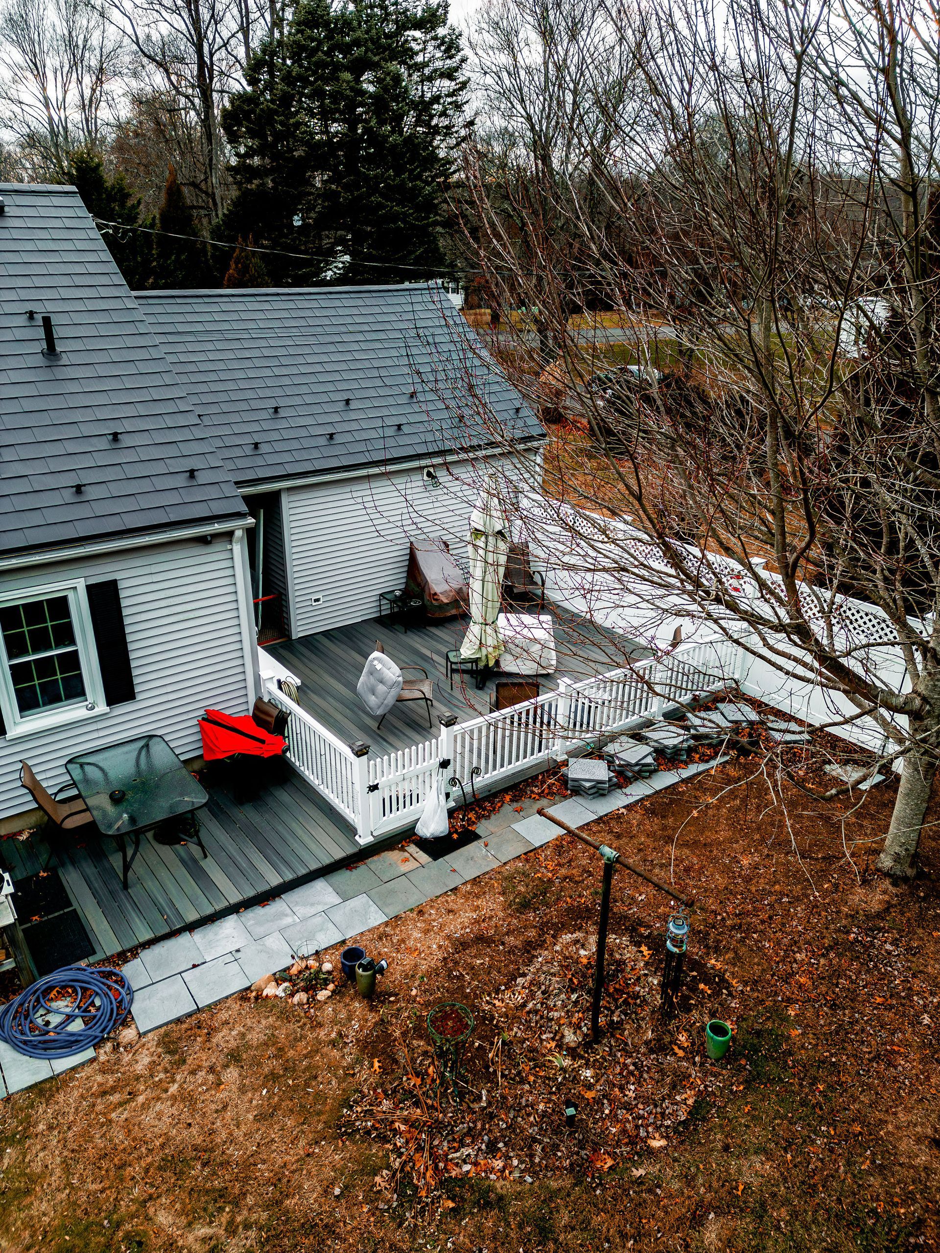 House with a black roof, white siding, and a wooden deck. Brown yard and bare trees surround.