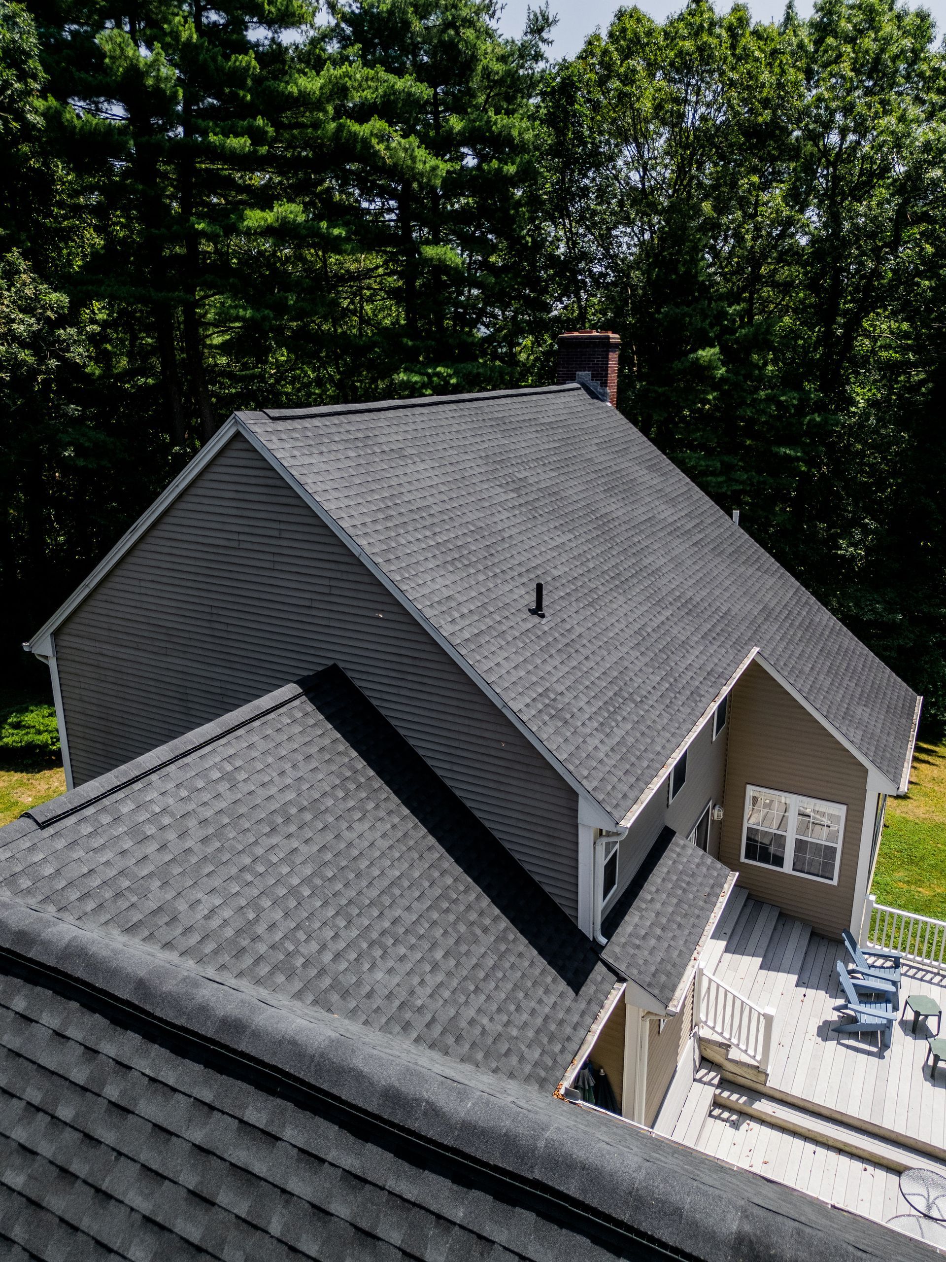 Dark gray shingled roof of a house with light brown siding, overlooking a deck with trees in the background.