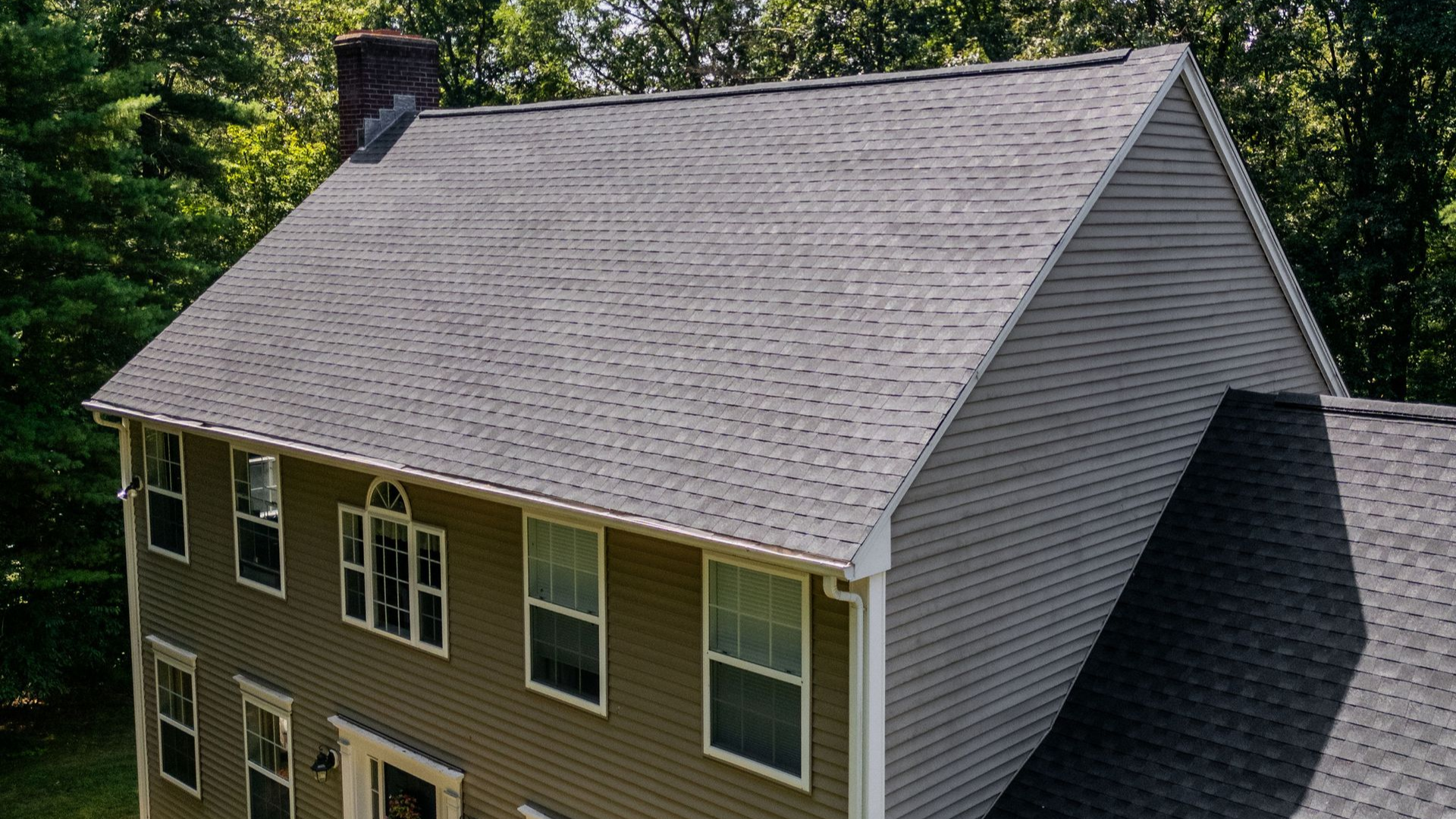 Two-story house with gray shingles and siding, windows, and a chimney against a green tree backdrop.