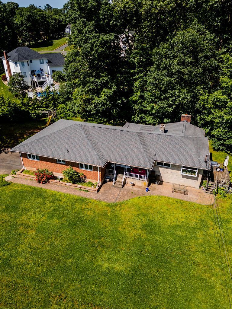 Aerial view of a single-story house with a gray roof and brick facade surrounded by green lawn and trees.