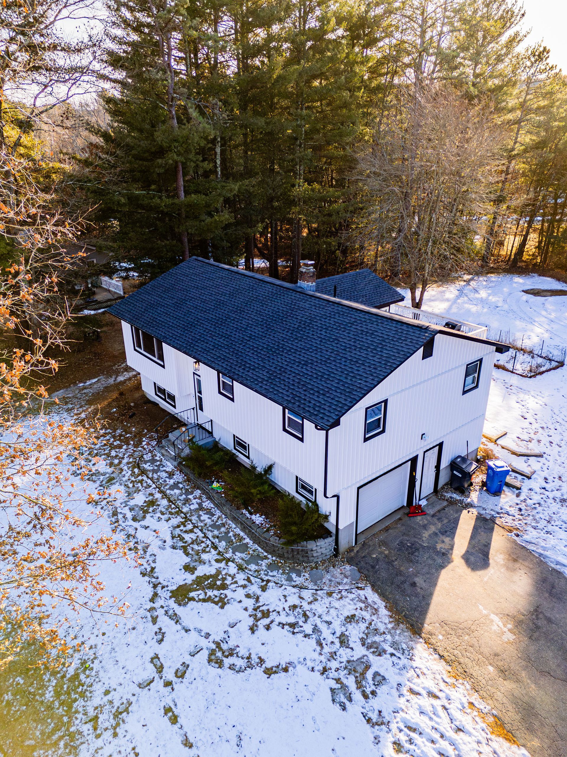 White house with a dark blue roof, snow on the ground, trees in the background, and a driveway.