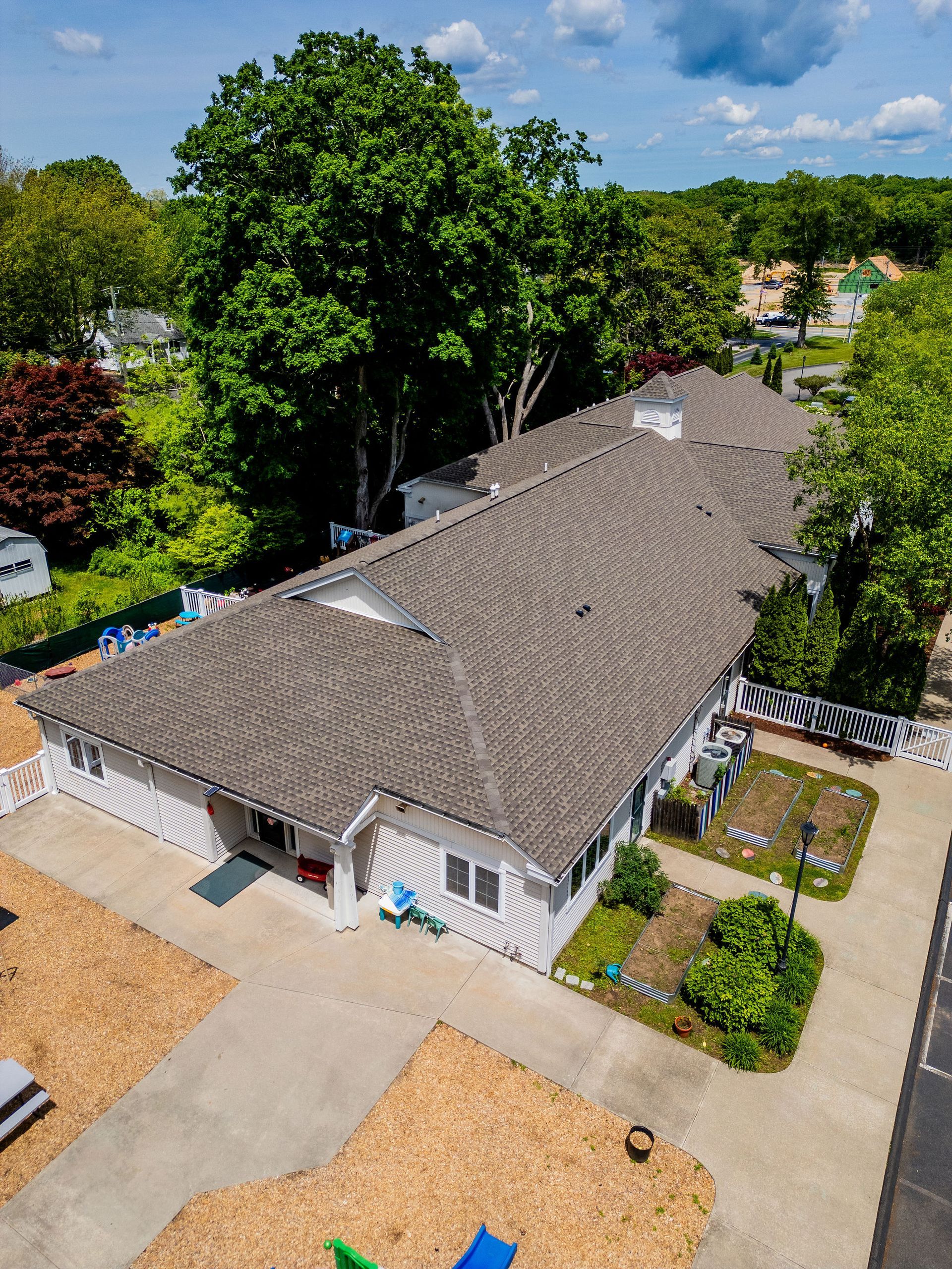 Aerial view of a white building with a gray roof, surrounded by trees and a concrete parking area.