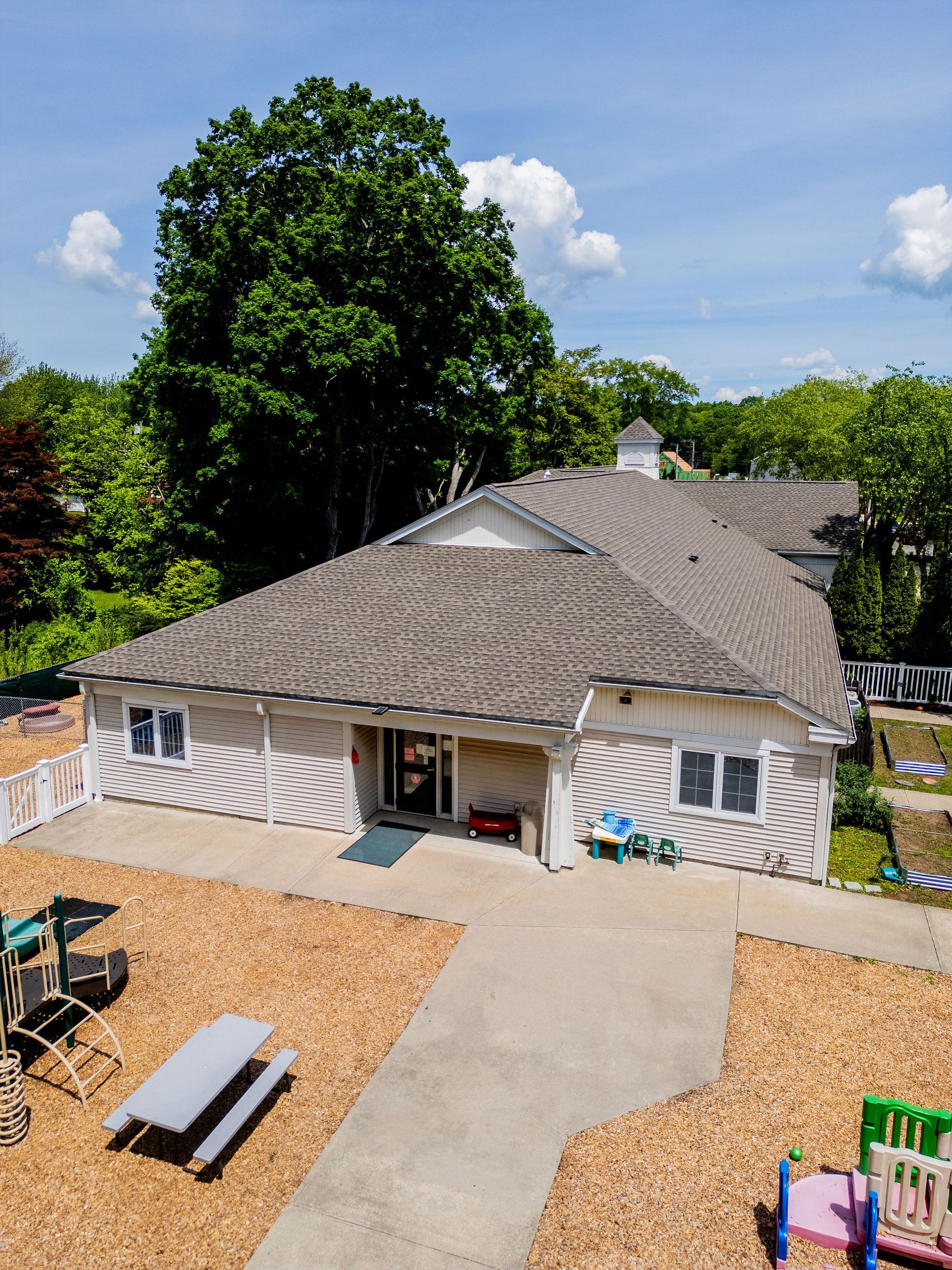 Daycare building with playground; concrete path, brown wood chips, green trees.