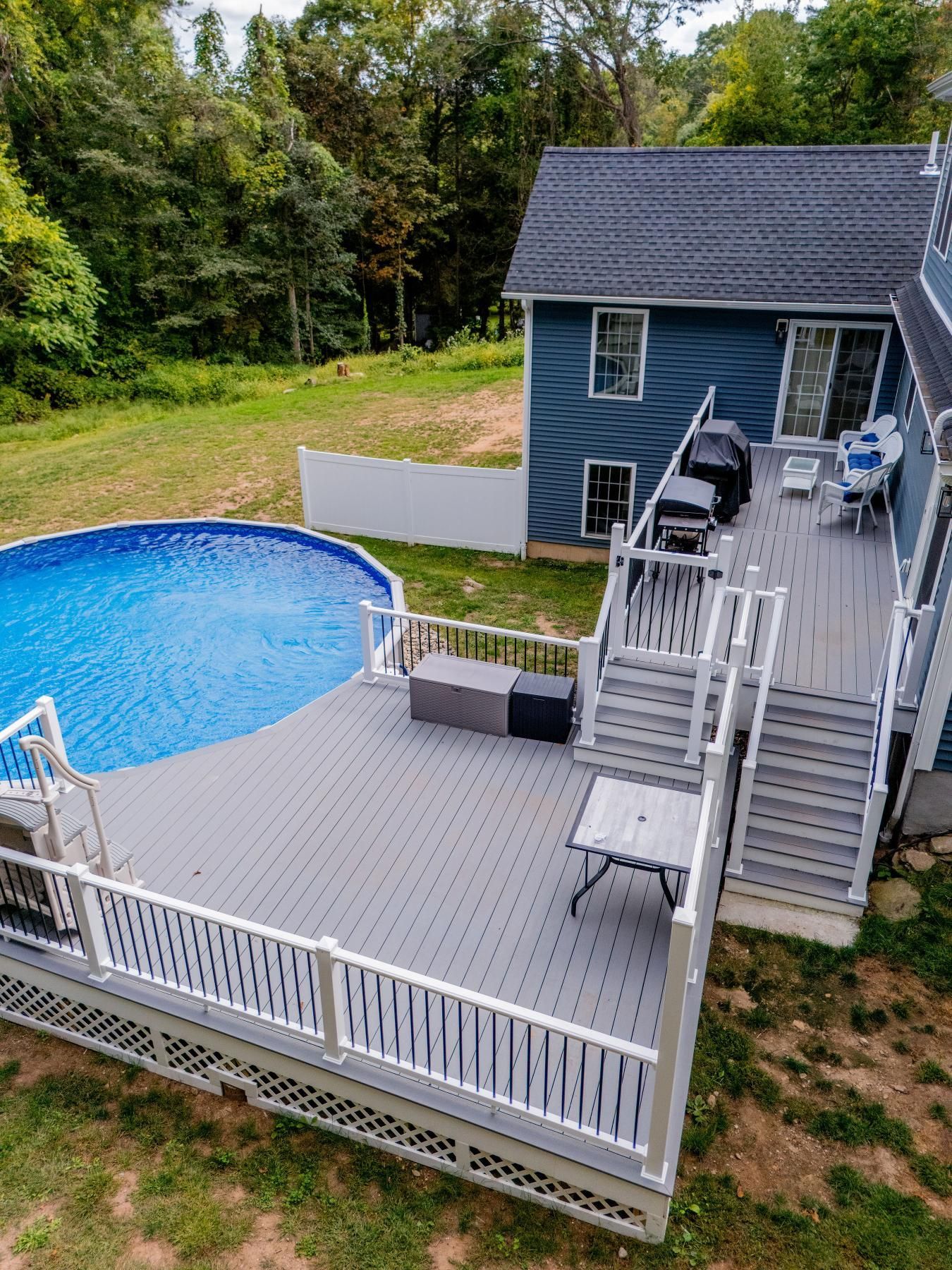 Deck with above-ground pool and stairs leading to a blue house with trees in the background.
