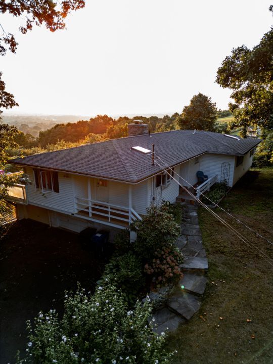 Single-story house on a hillside at sunset; white siding, dark roof, small porch, and stone steps leading to the house.