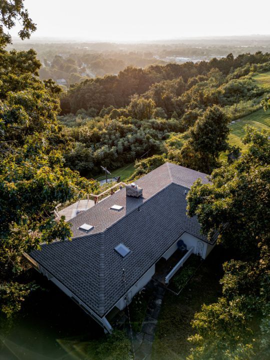 Overhead view of a house with a dark roof surrounded by green trees, with a distant, bright sunset.
