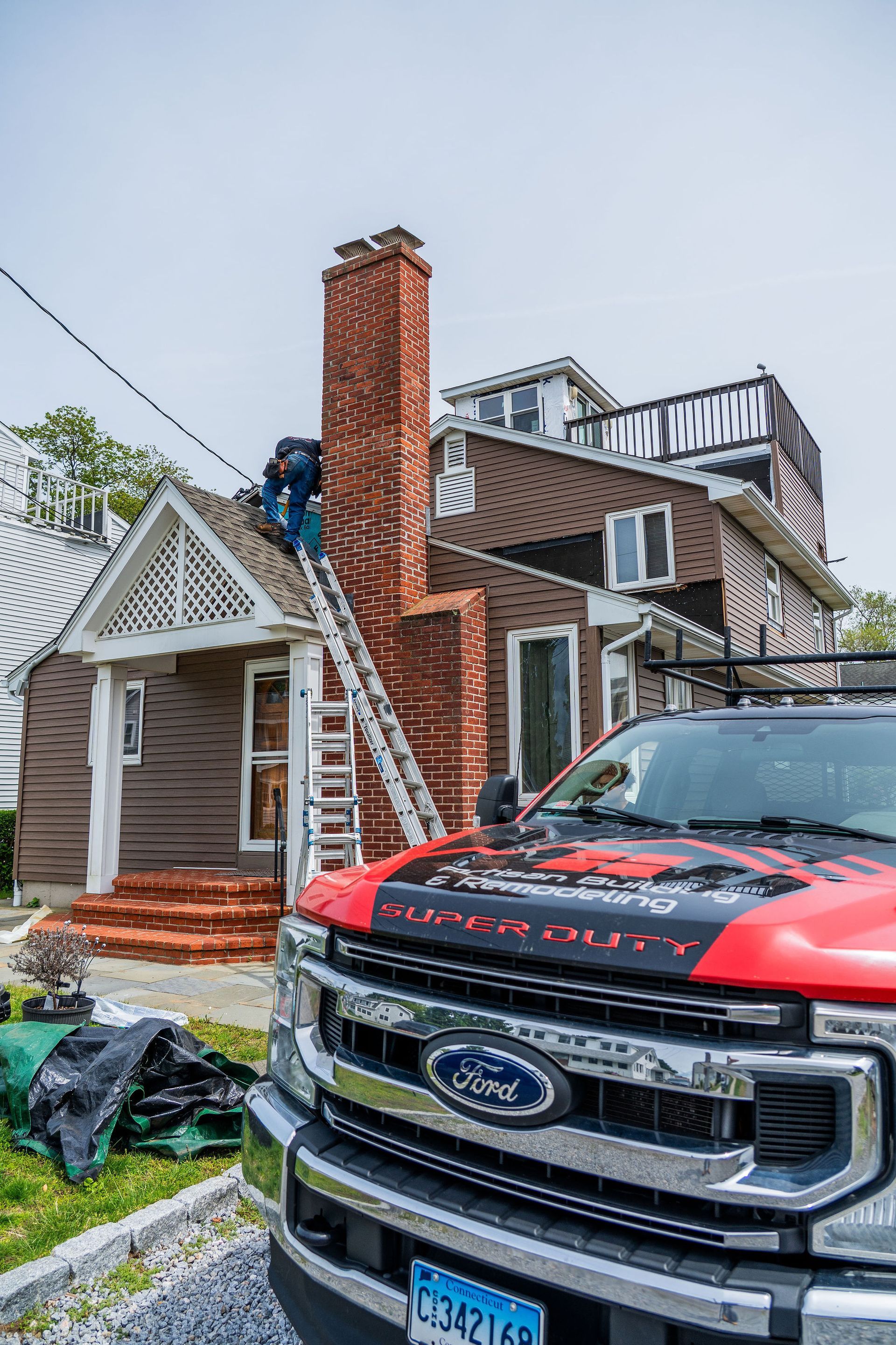 A worker on a ladder repairs a brick chimney on a brown house. A red Ford truck is in the foreground.