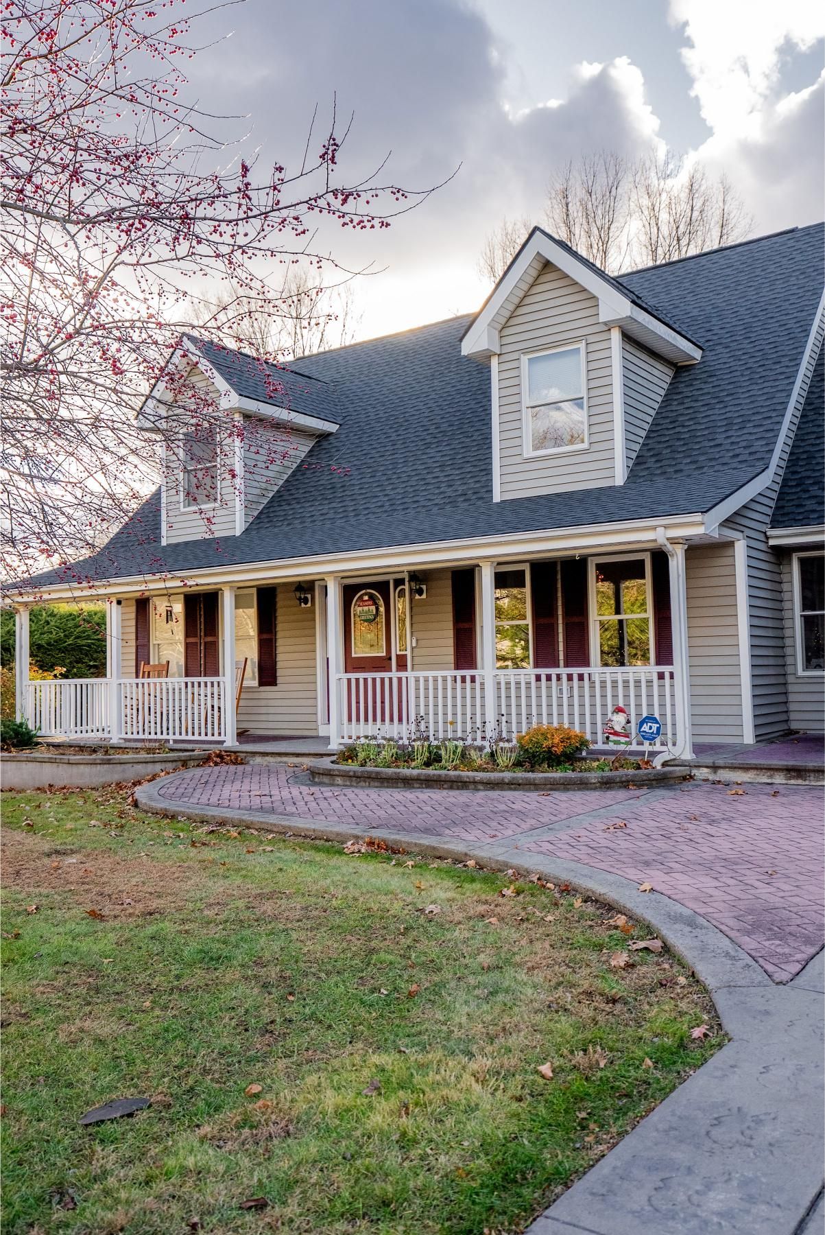 Beige house with a dark roof, porch, and a brick driveway under a cloudy sky.