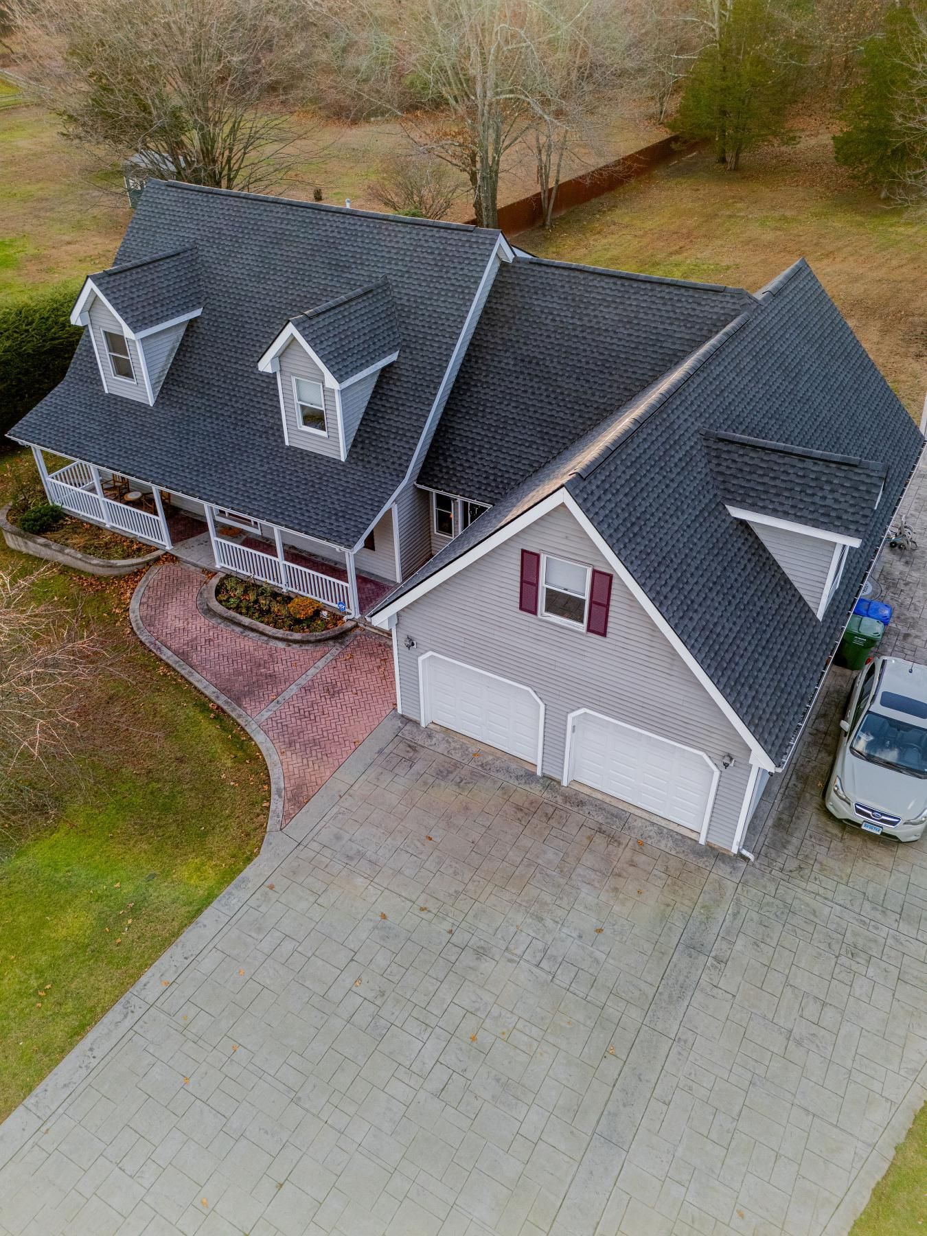 Gray house with dark roof and two-car garage; brick driveway, green yard, car parked outside.