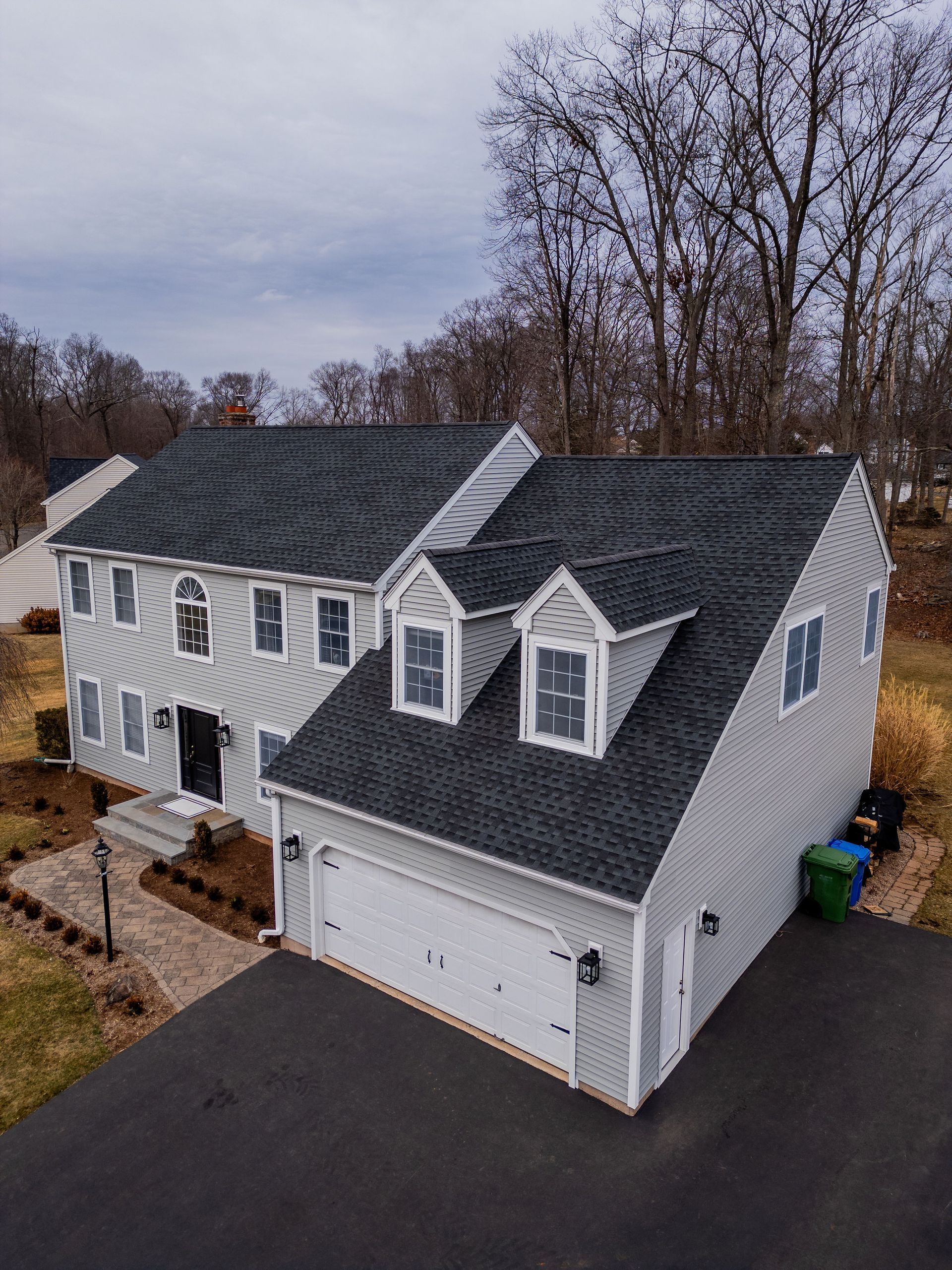 Gray two-story house with a black asphalt driveway, garage, and two dormers. Overcast sky.