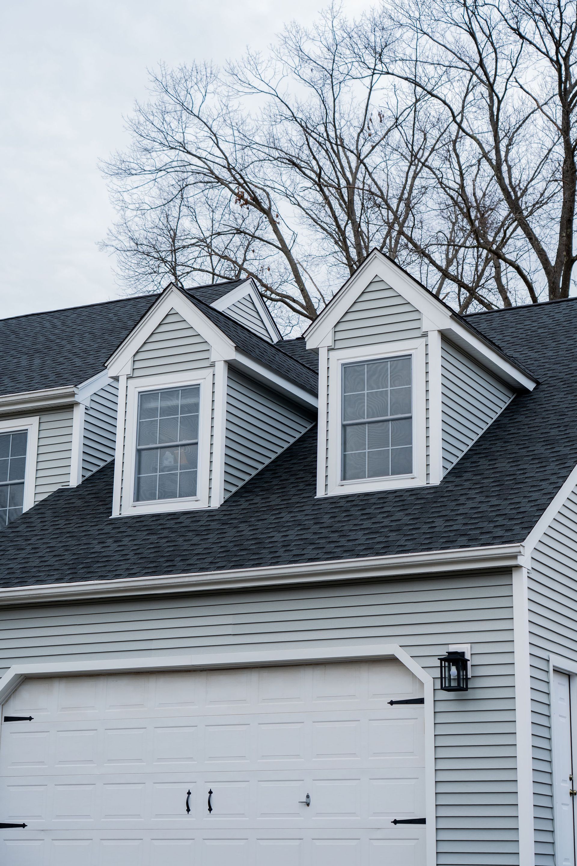 Gray house with white garage door and dormer windows under a cloudy sky.