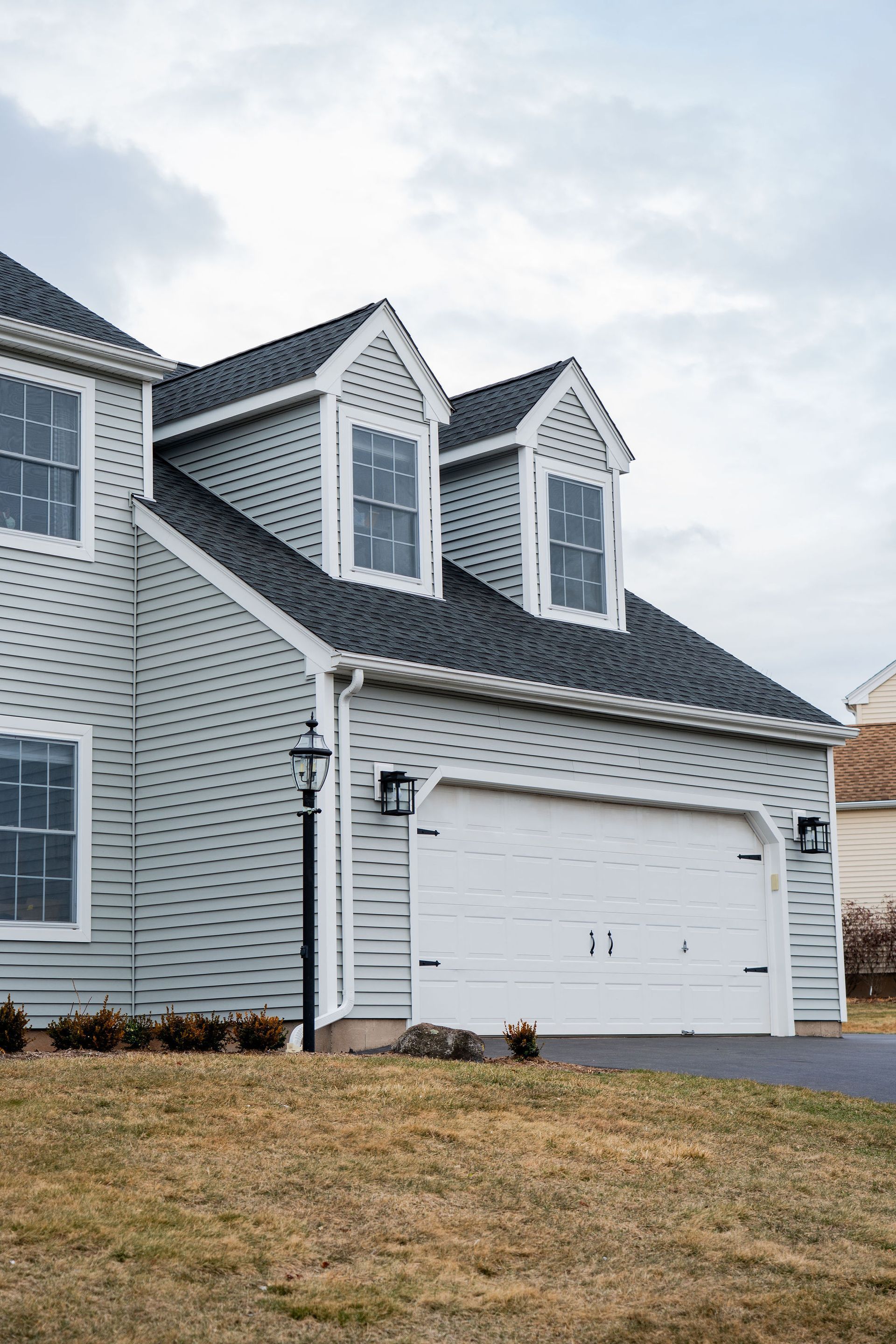 Gray house with white garage door and dormer windows under cloudy sky.