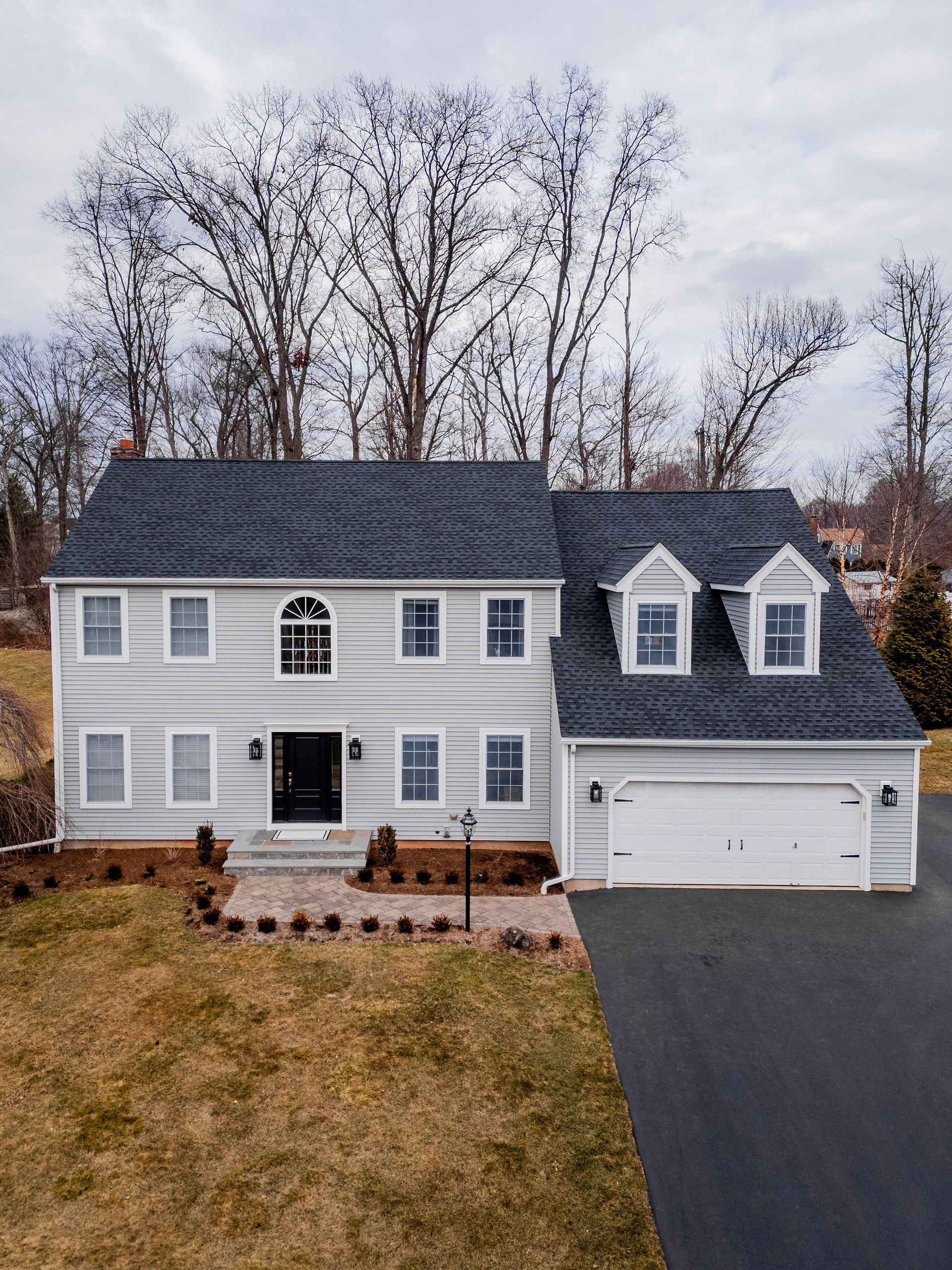Two-story light gray house with black roof, white trim, and a two-car garage. A long, black driveway leads up to it.