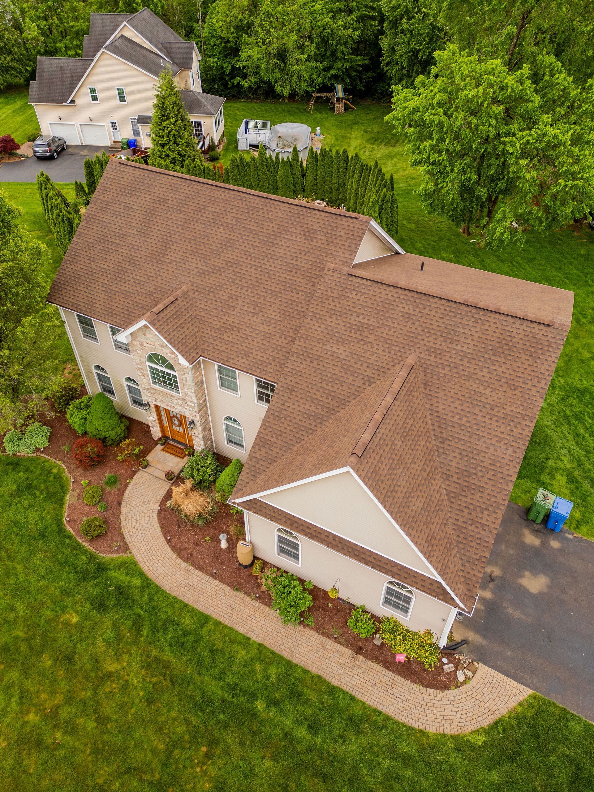 Aerial view of a two-story beige house with a brown roof and a curved stone walkway surrounded by green grass.