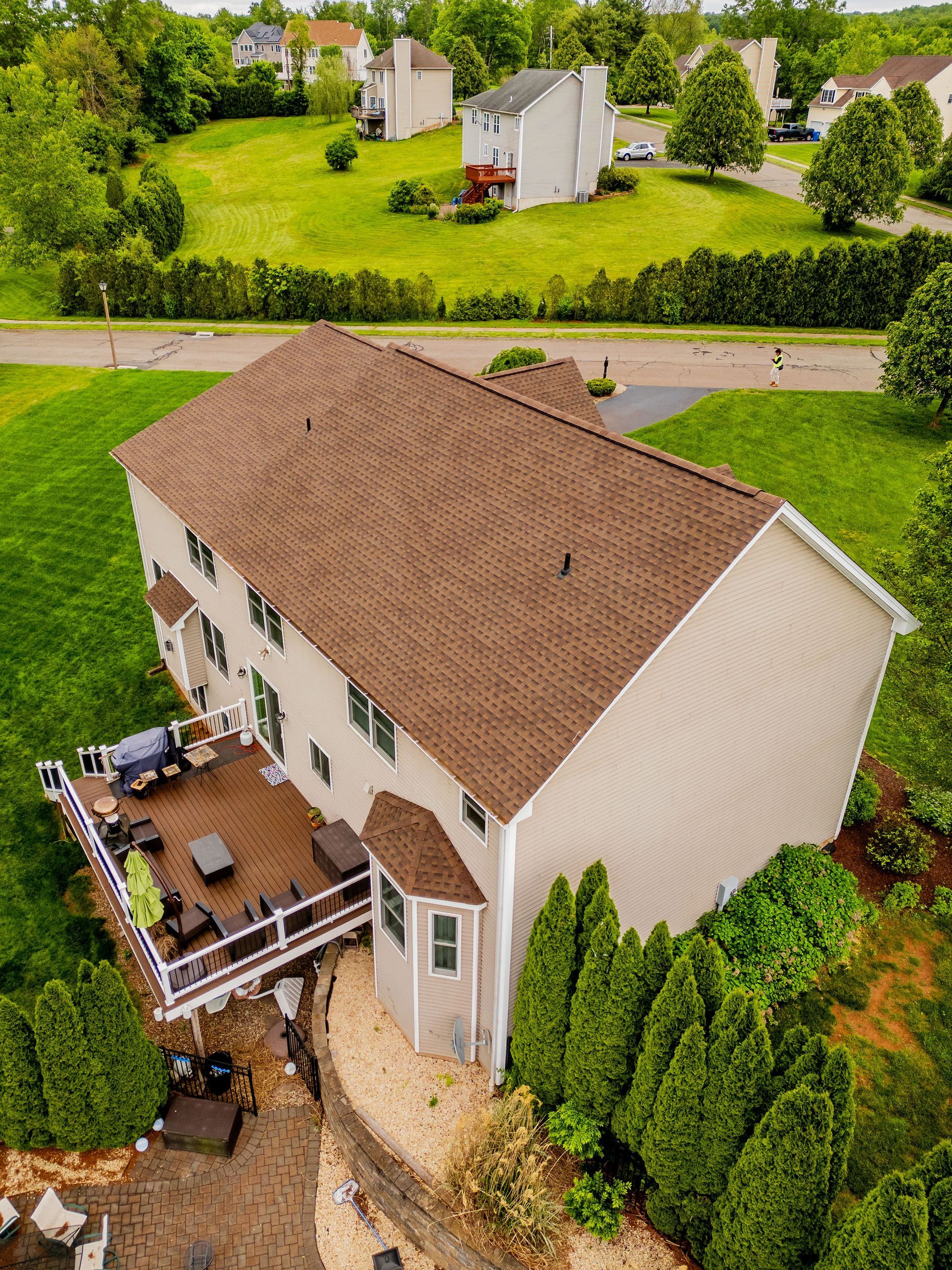 Aerial view of a two-story beige house with a brown roof and a wooden deck overlooking a green yard and neighborhood.