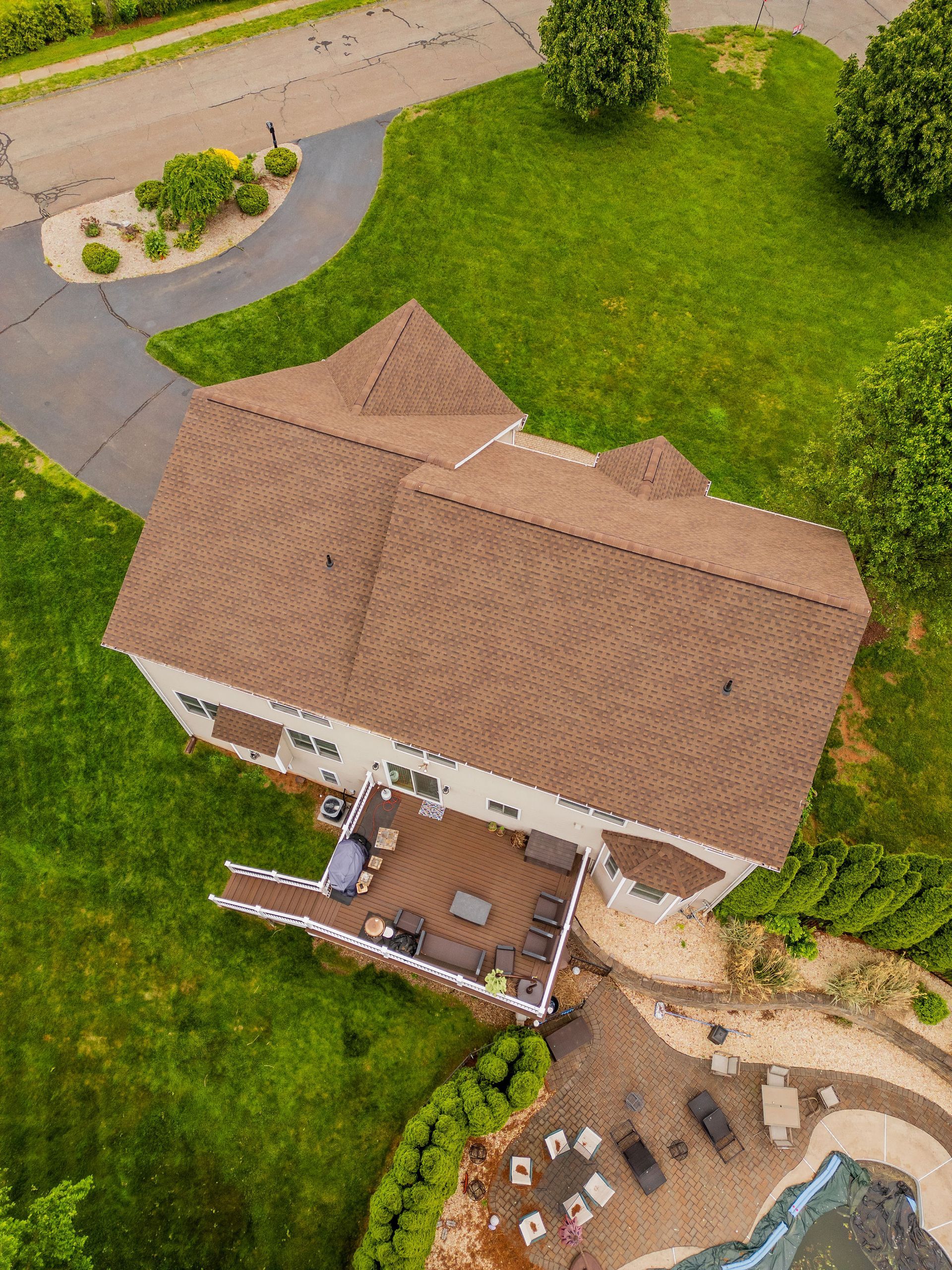 Aerial view of a house with a brown roof and a deck, surrounded by green grass, trees, and a driveway.
