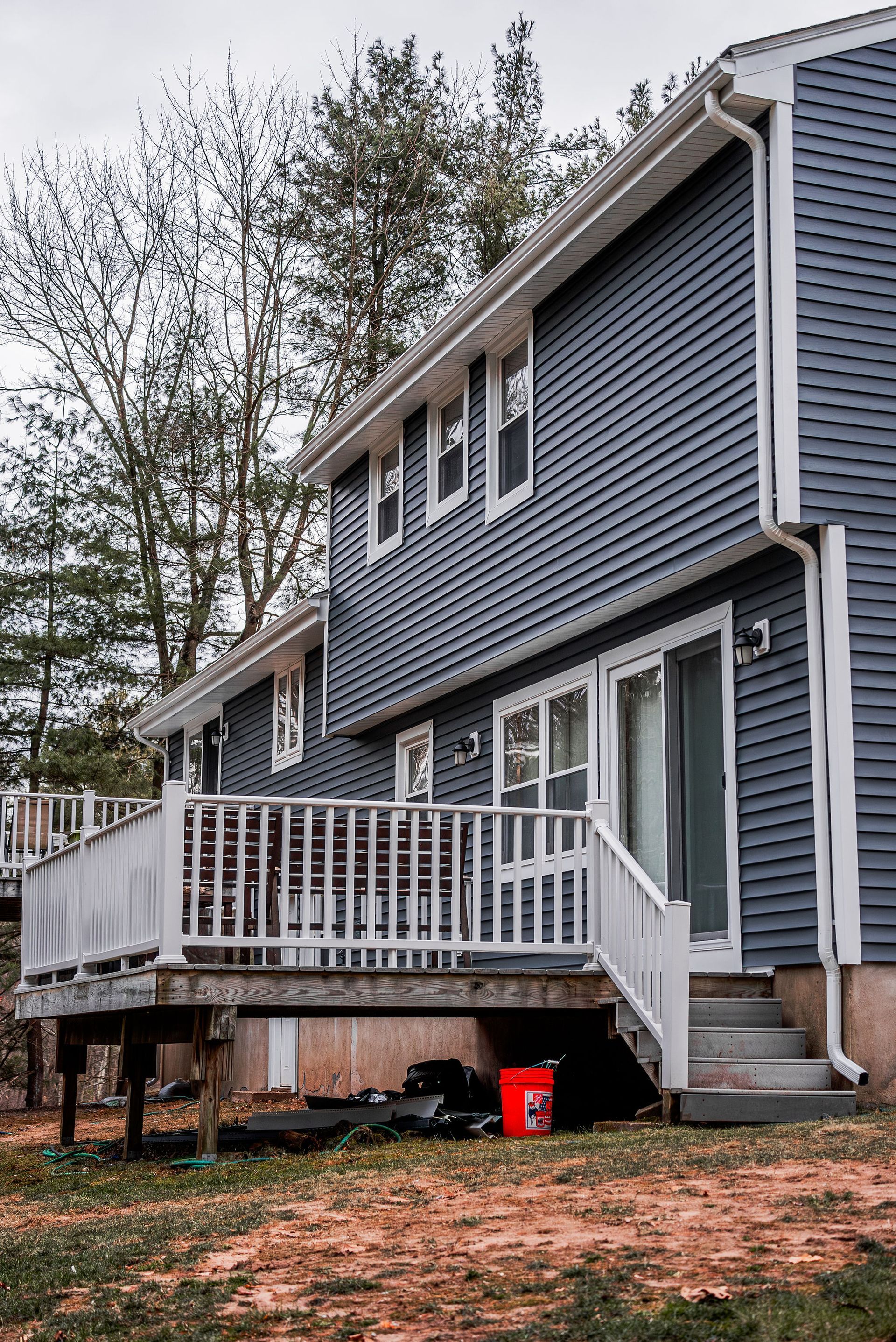 Two-story house with blue siding, white deck, and a red bucket on the ground.