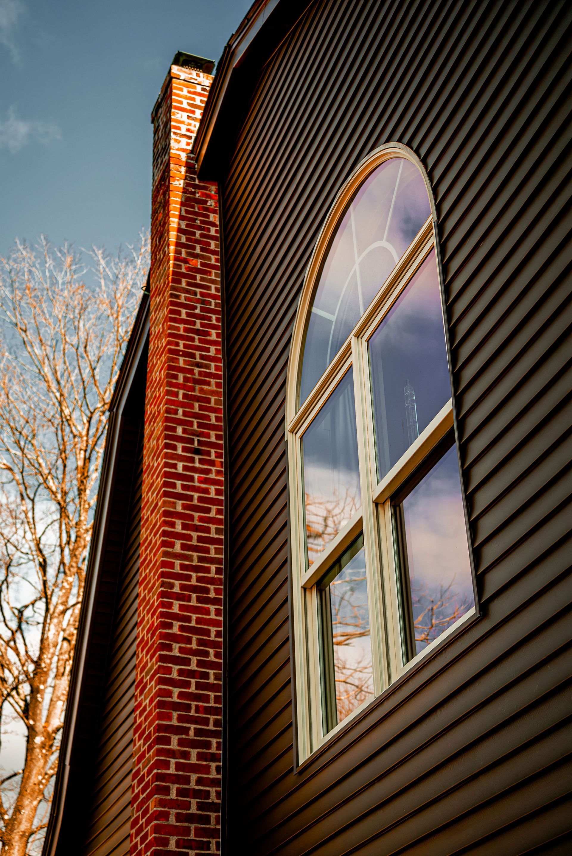 Brick chimney next to a dark-sided building with arched window reflecting the sky and bare tree.