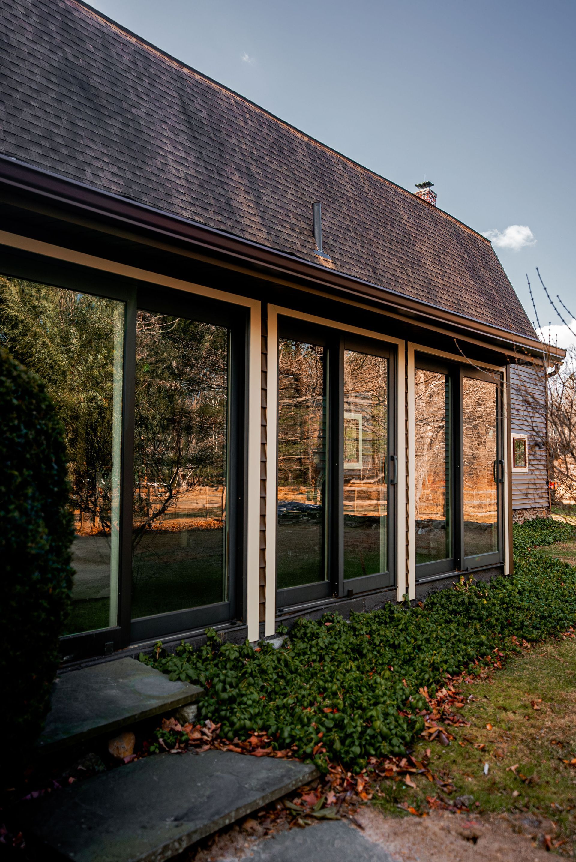 Side of house with large windows reflecting trees and sky, surrounded by greenery. Brown roof.