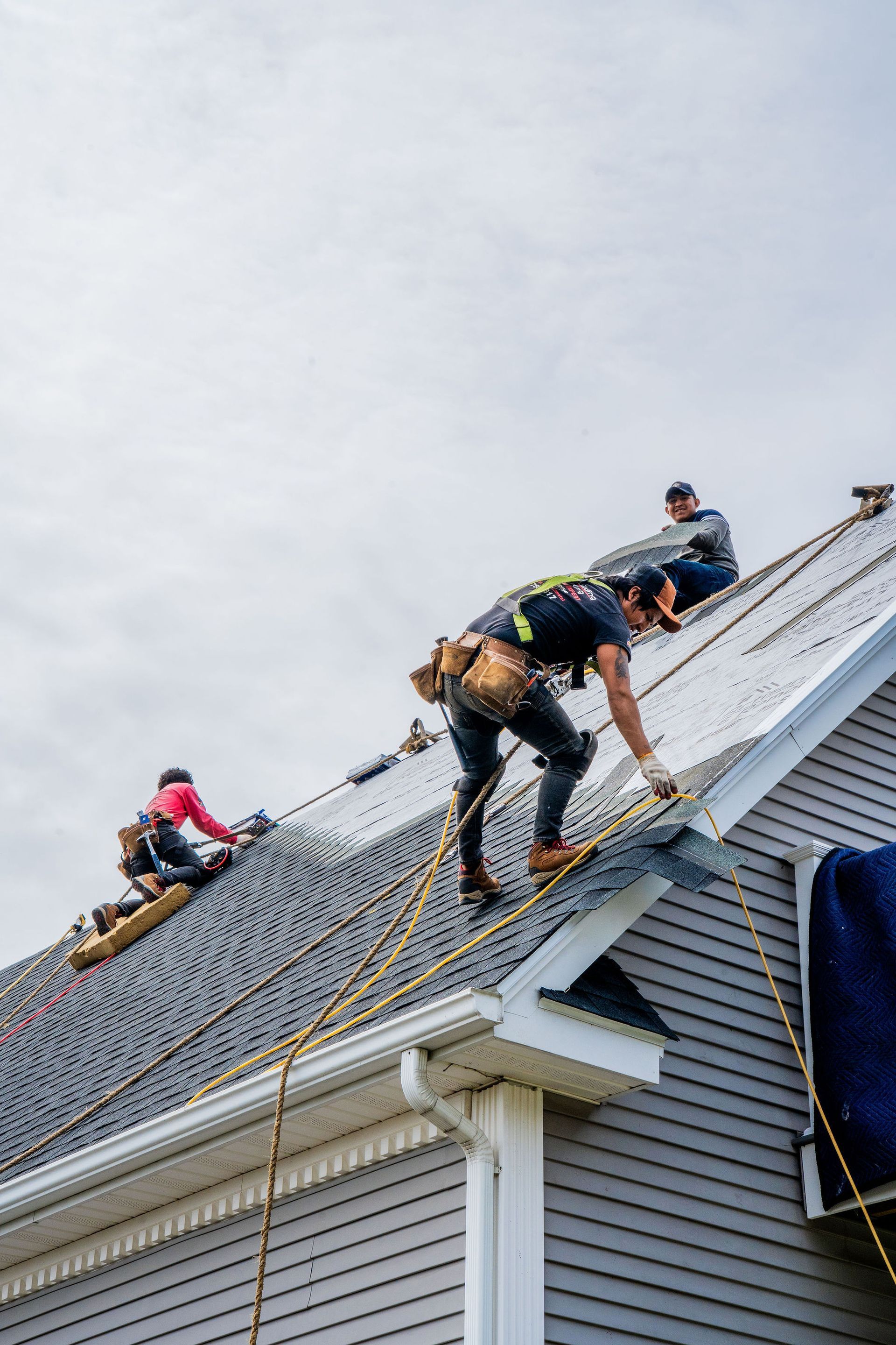 Roofers working on a house roof under a cloudy sky, using safety harnesses.