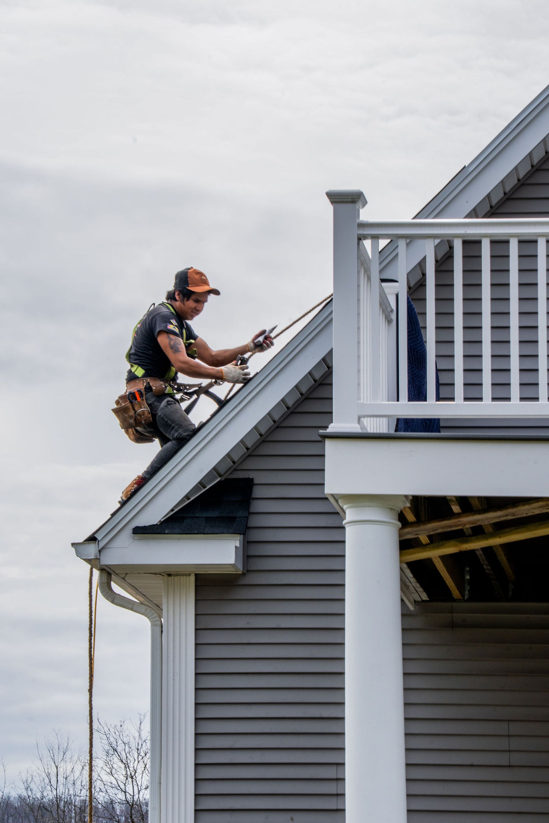 Construction worker on a roof, secured with ropes. Gray siding, white trim, cloudy sky.