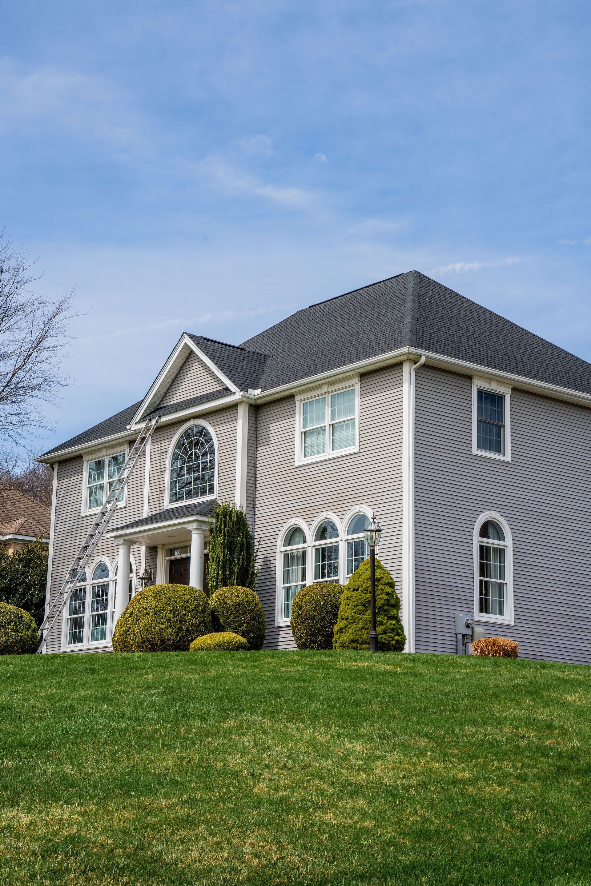 Two-story gray brick house with arched windows, nestled on a grassy hill, under a blue sky.