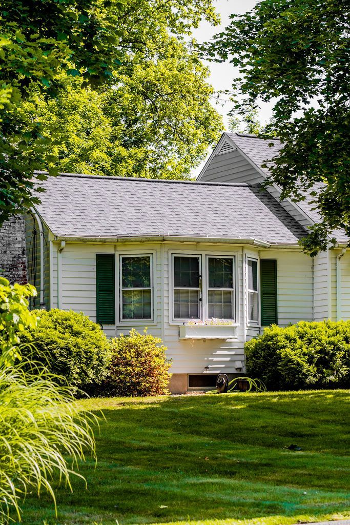 White house with green shutters, gray roof, and green shrubbery on a sunny day.