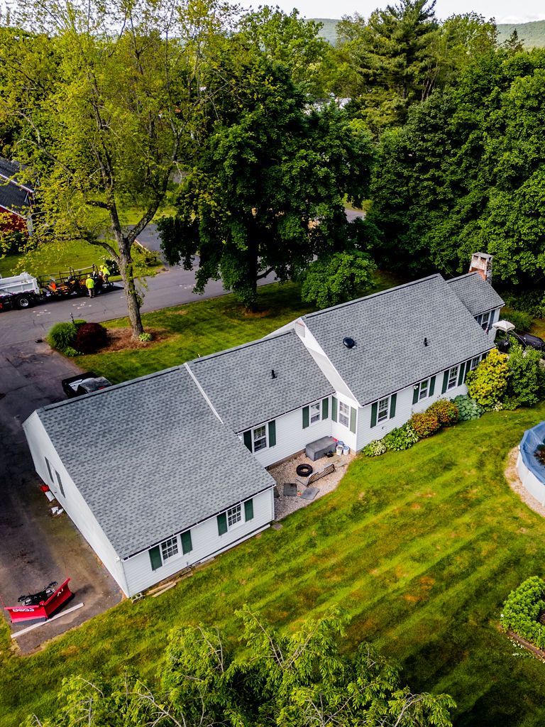 Aerial view of white house with gray roof, green lawn, trees, and road.