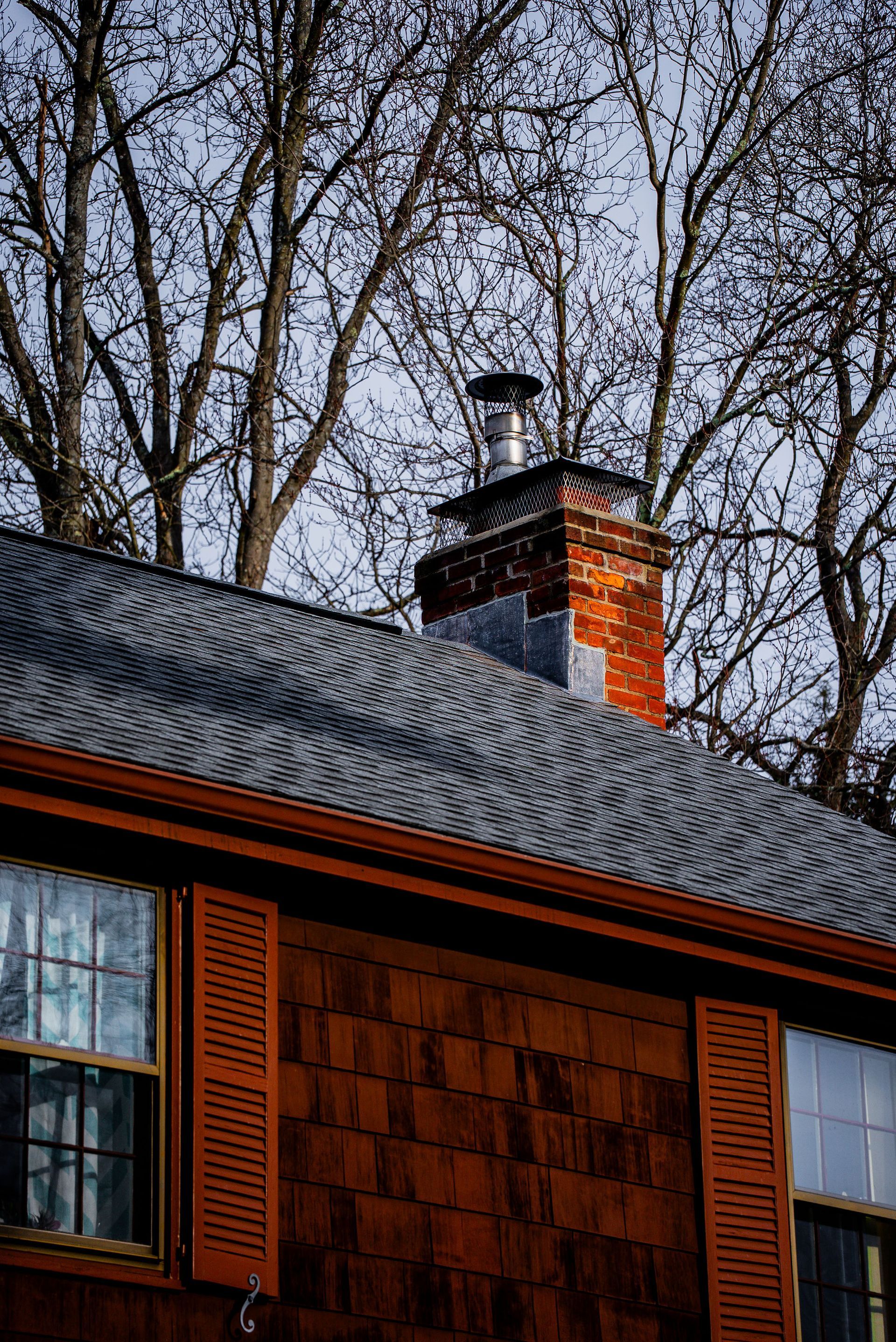 Brick chimney on a brown-shingled house with wooden shutters, bare tree branches in the background.